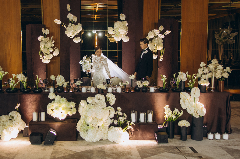 Bride and groom walking arm-in-arm down a white aisle decorated with tall vases of white flowers at an outdoor wedding ceremony.