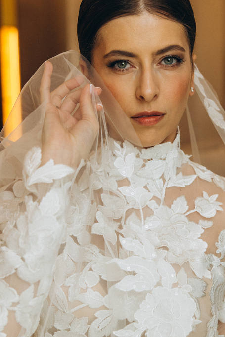 Woman in an elegant backless wedding dress standing by a window with sheer curtains looking outside at greenery.