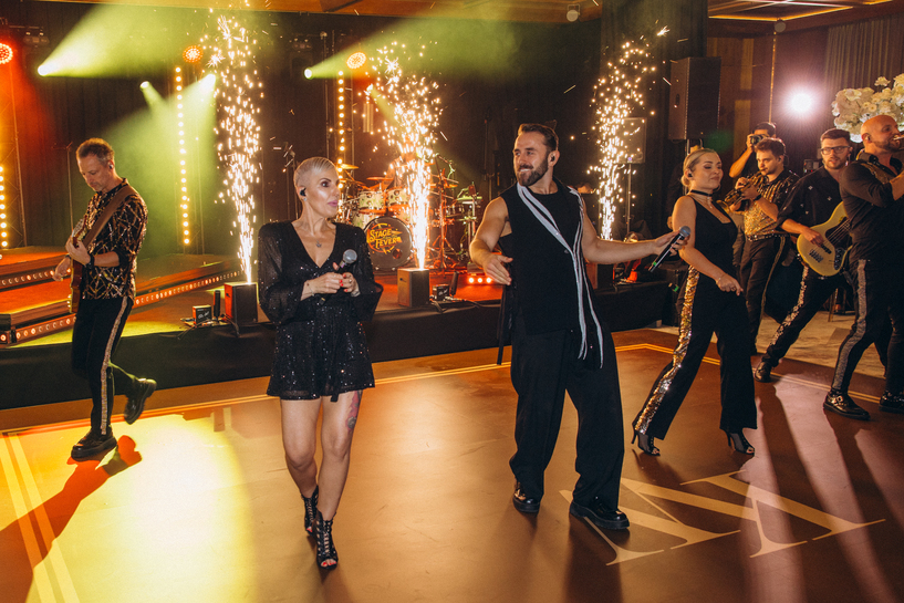 Couple dancing on a patterned lit floor, the woman in a white lace wedding dress and the man in a dark suit.