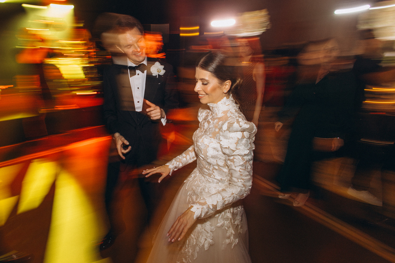 Couple dancing on a patterned lit floor, the woman in a white lace wedding dress and the man in a dark suit.