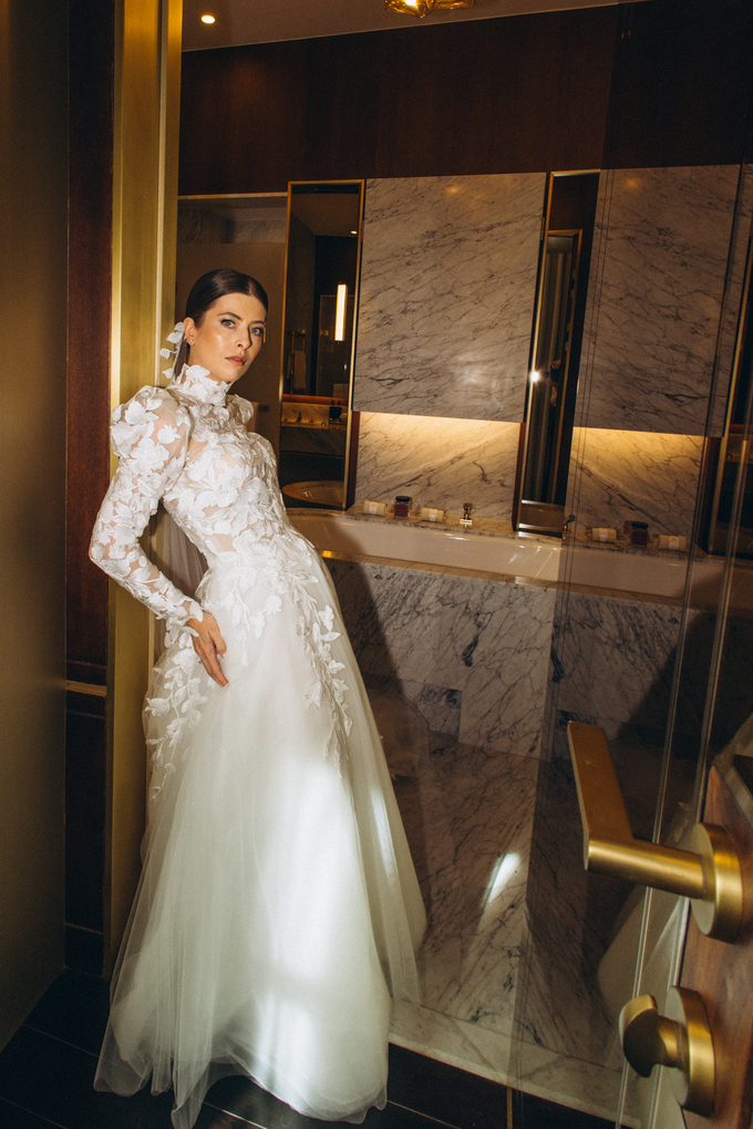 Woman in an elegant backless wedding dress standing by a window with sheer curtains looking outside at greenery.