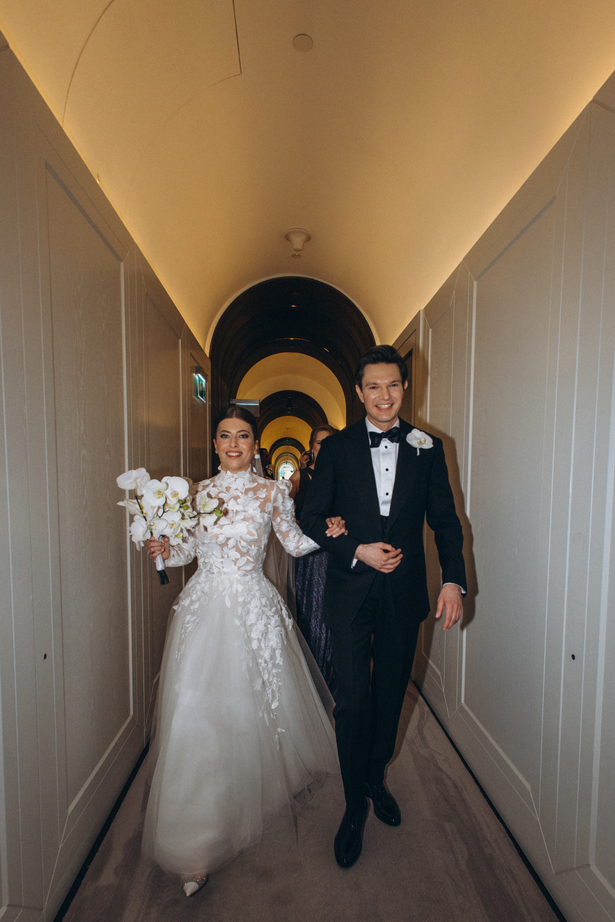 Bride and groom walking arm-in-arm down a white aisle decorated with tall vases of white flowers at an outdoor wedding ceremony.