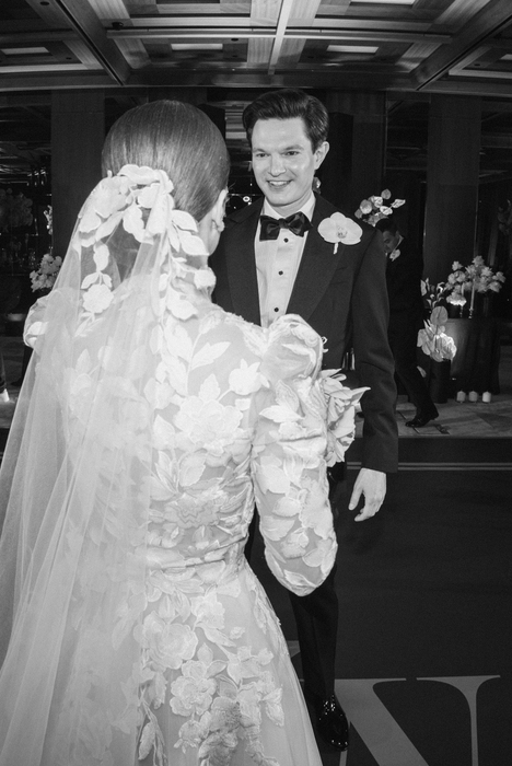 Woman in an elegant backless wedding dress standing by a window with sheer curtains looking outside at greenery.