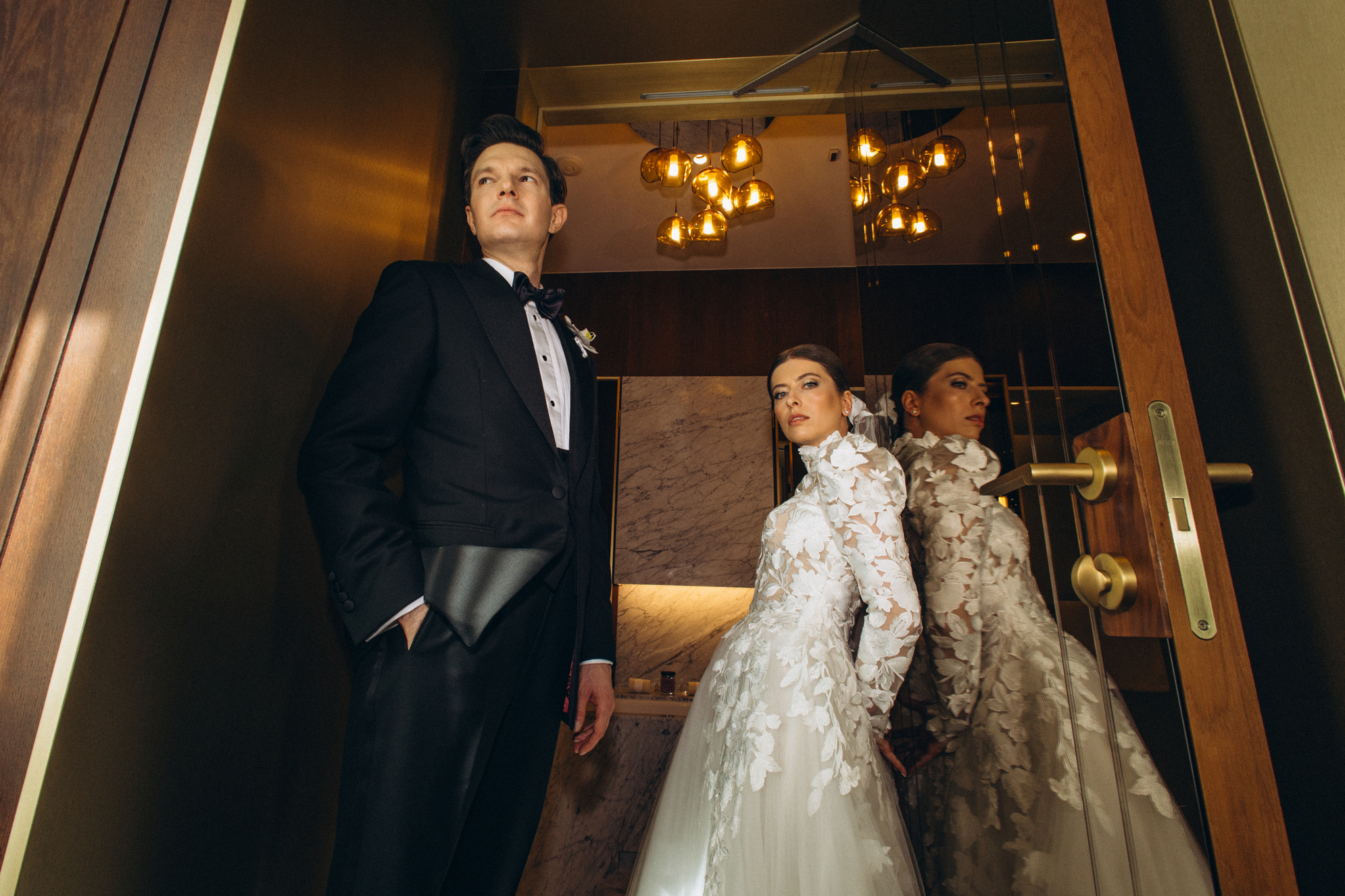 Woman in a backless lace wedding dress standing by a bright window with sheer curtains.