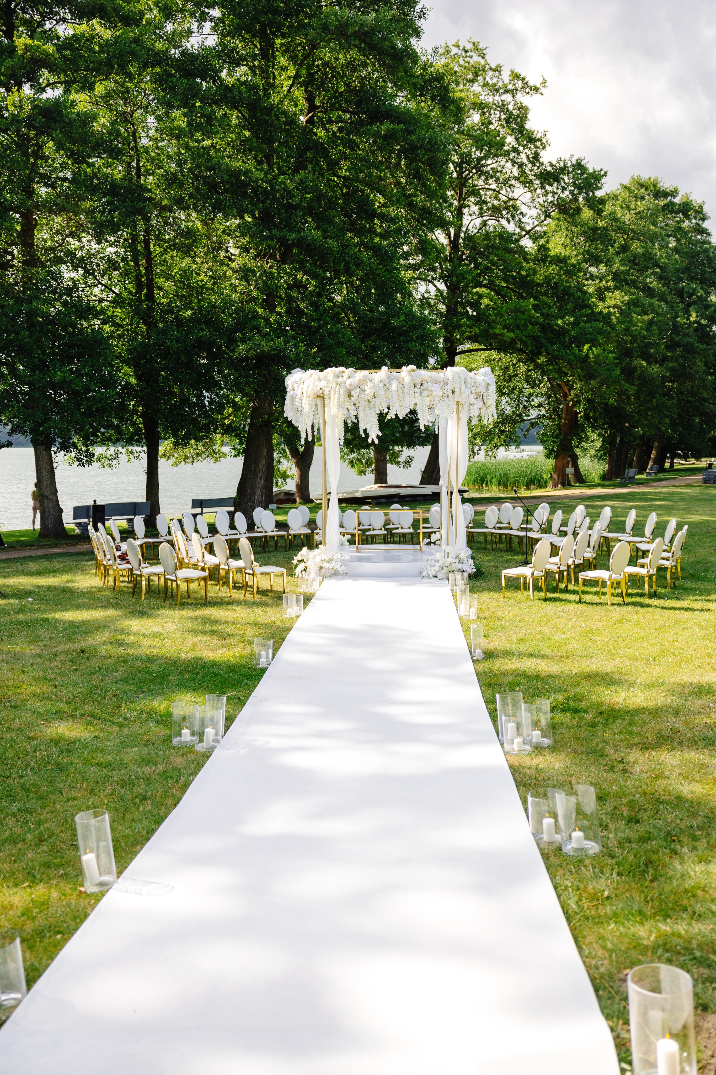 Smiling woman in an off-shoulder white dress with jewellery, standing on a sunlit pier with a man blurred in the background.