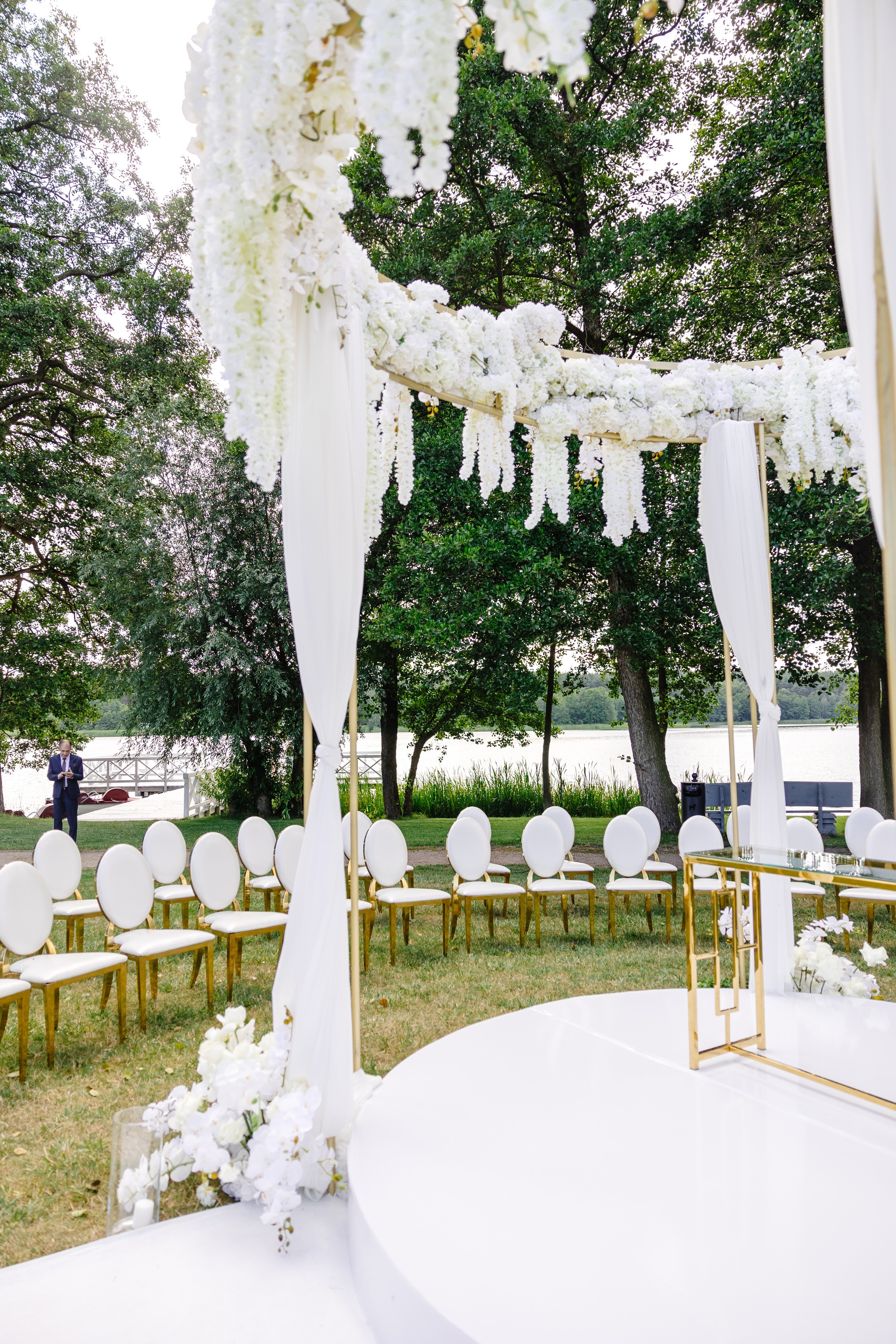 Couple dressed in white standing on a wooden pier overlooking a calm lake surrounded by trees at sunset.