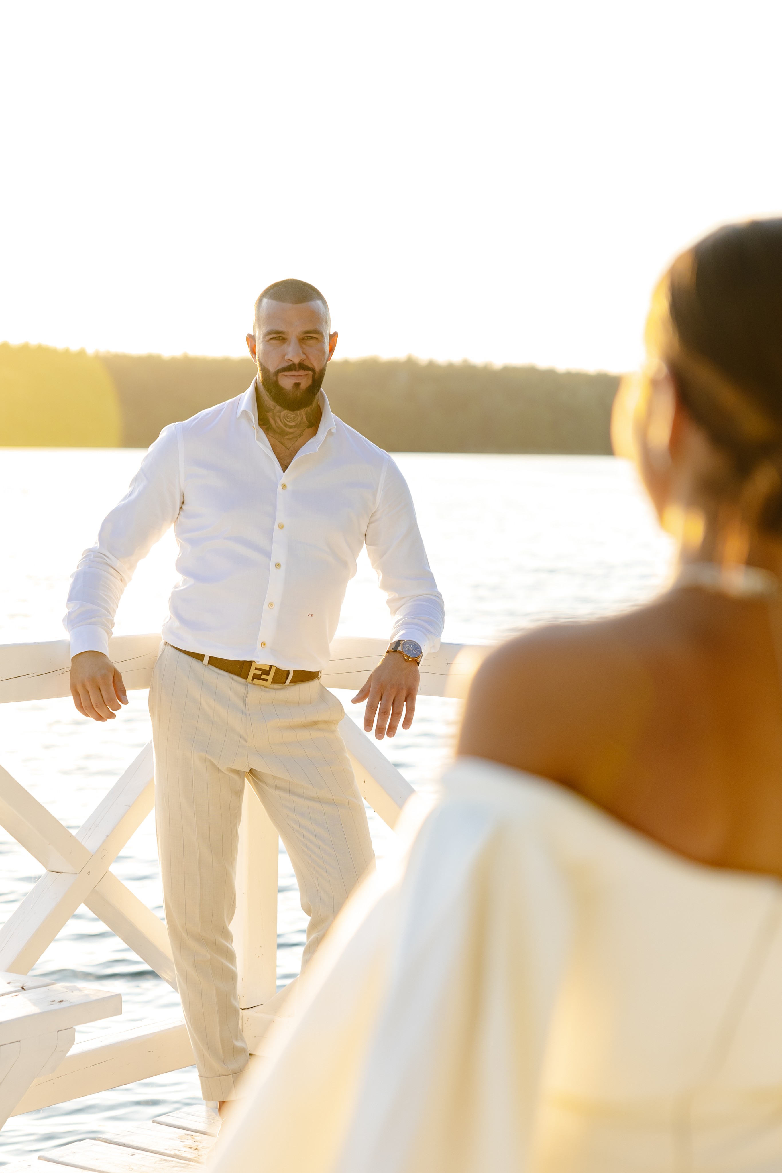 Couple dressed in white standing on a wooden pier overlooking a calm lake surrounded by trees at sunset.