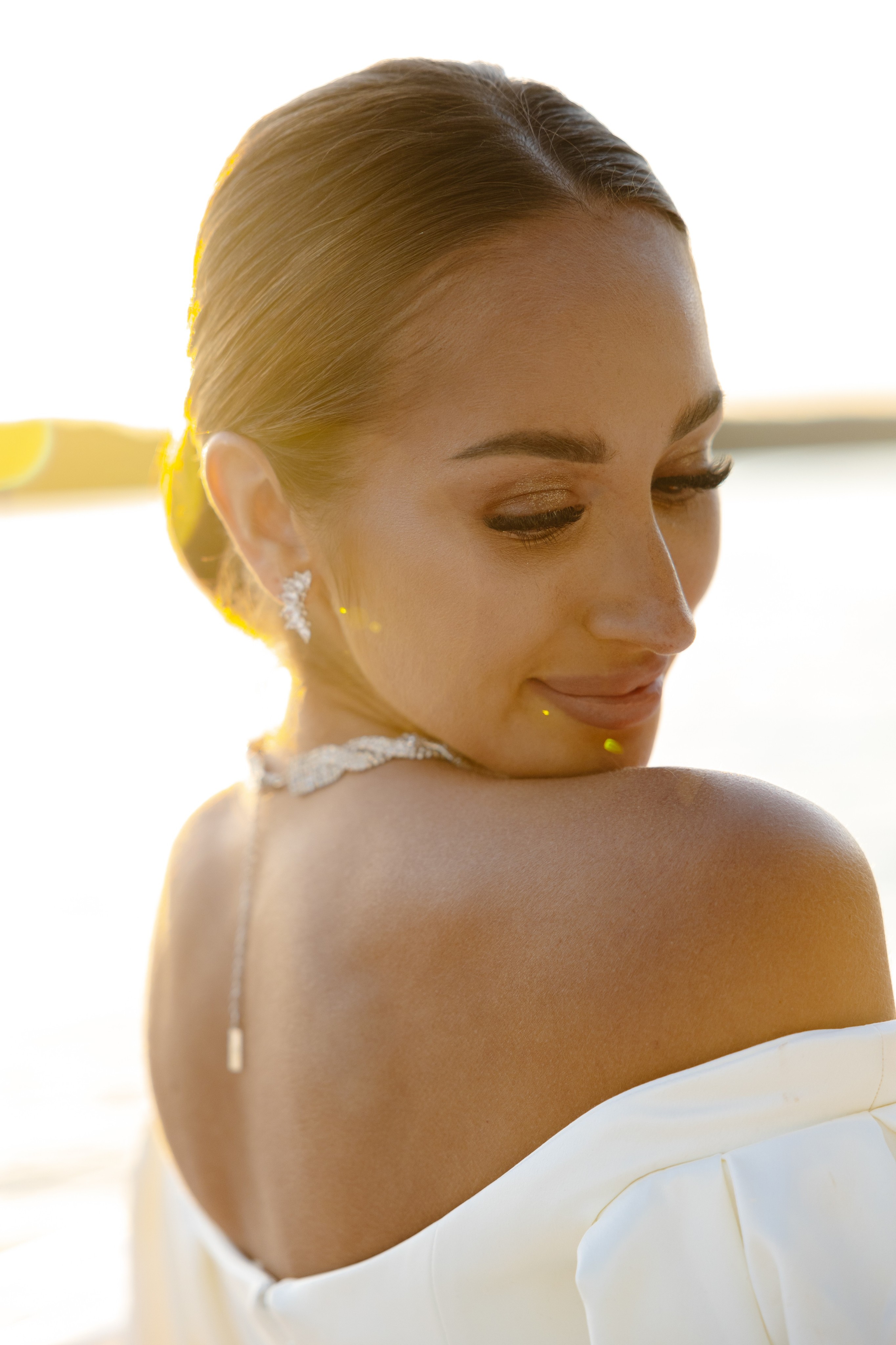 Couple dressed in white standing on a wooden pier overlooking a calm lake surrounded by trees at sunset.