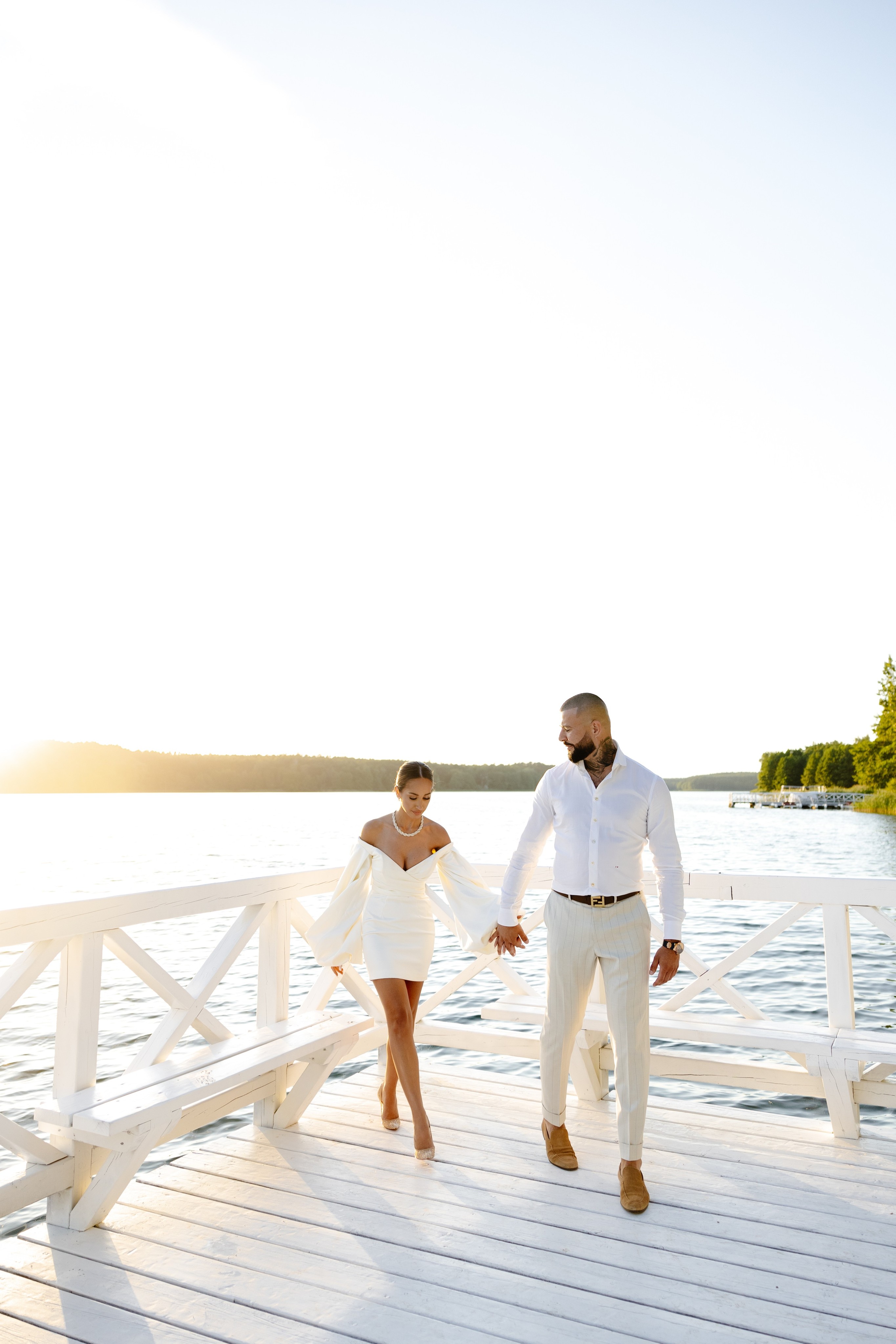 Couple dressed in white standing on a wooden pier overlooking a calm lake surrounded by trees at sunset.