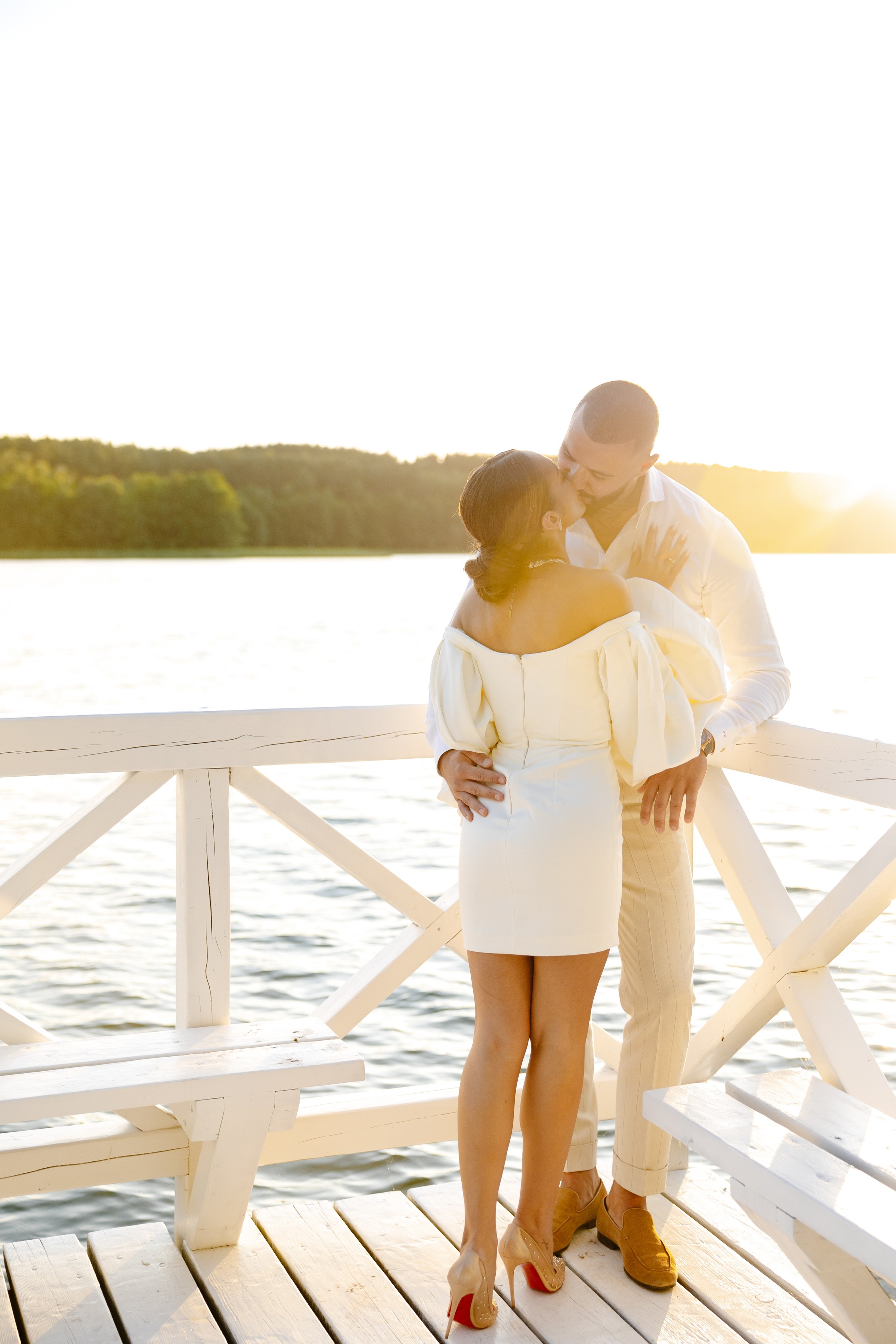 Couple dressed in white standing on a wooden pier overlooking a calm lake surrounded by trees at sunset.