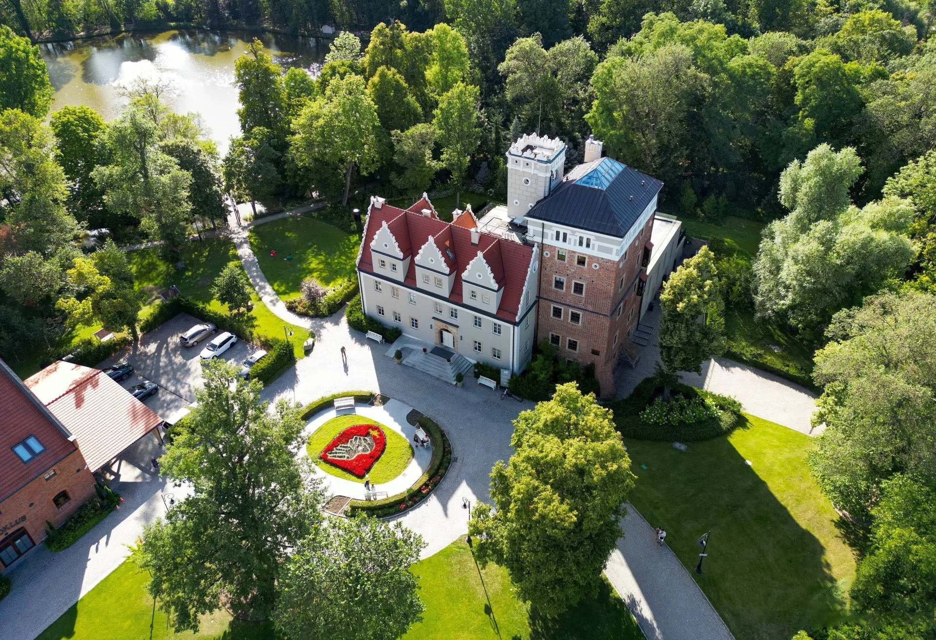 Aerial view of a historic building with red roof and tower surrounded by lush green trees, gardens, and a pond.