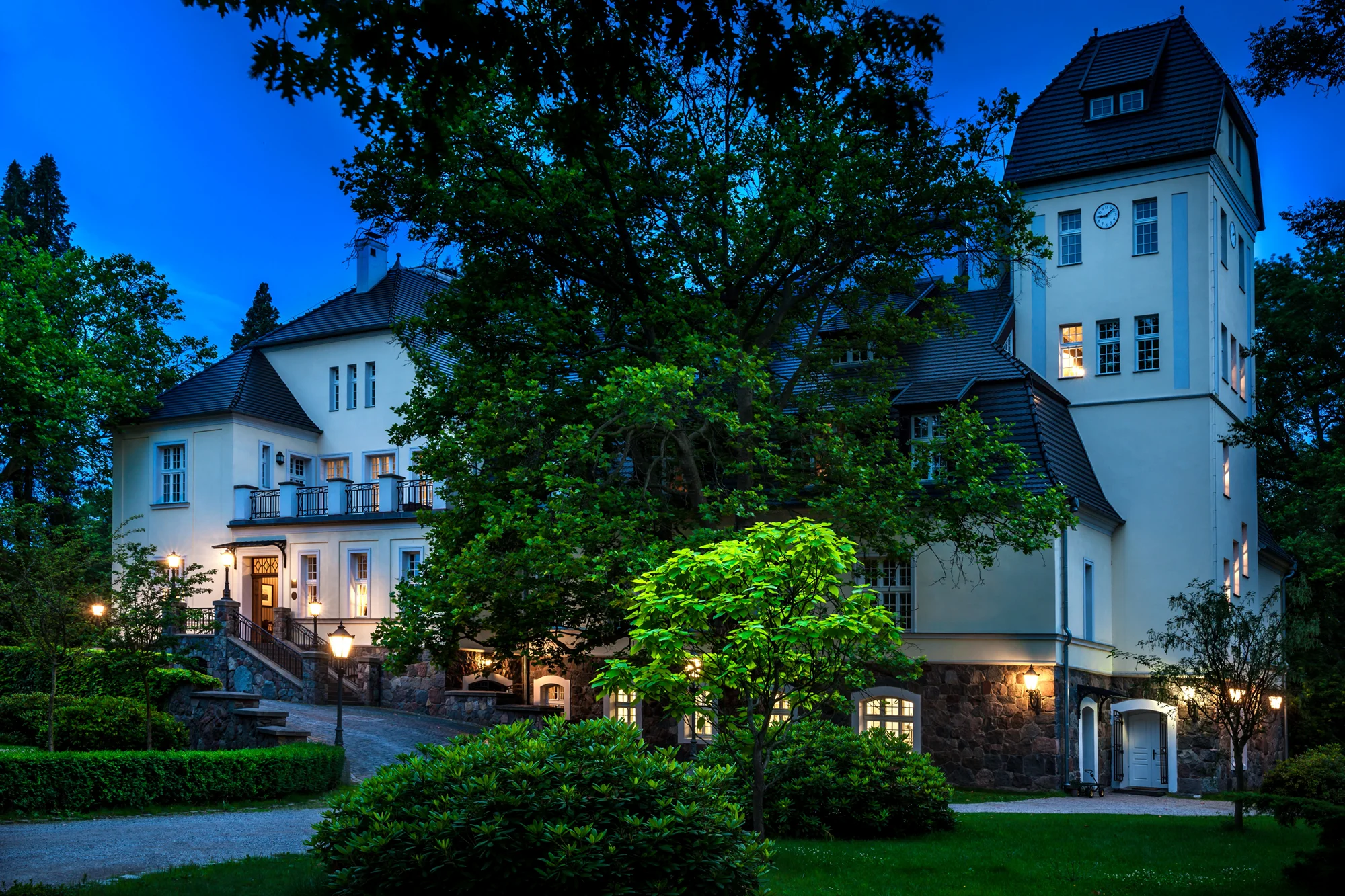Large historic mansion with lit windows at dusk surrounded by lush green trees and bushes.