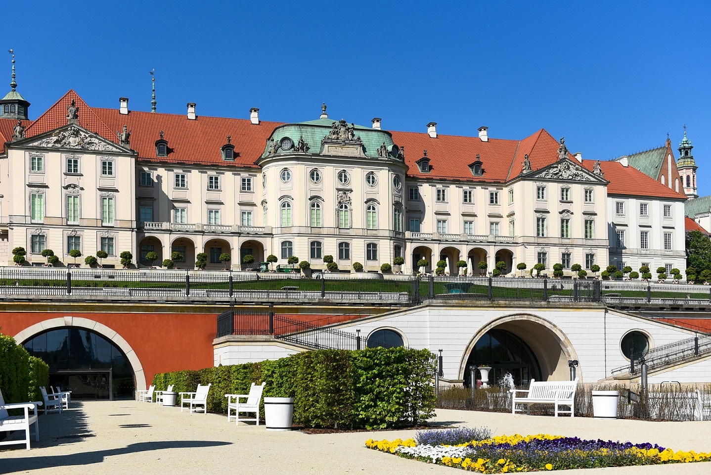 Historic European palace with red-tiled roof, ornate facade, and manicured gardens with white benches under a clear blue sky.