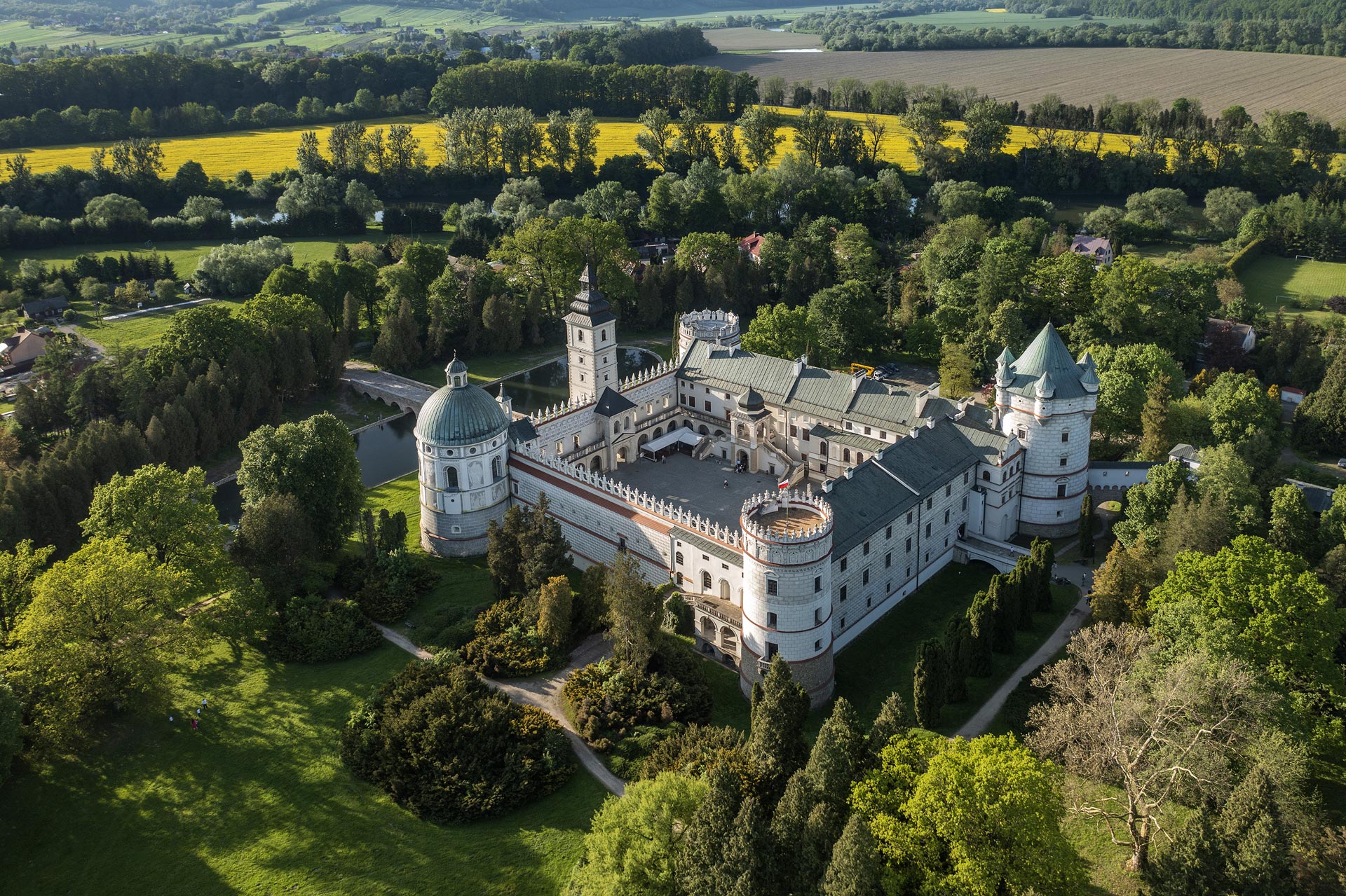 Aerial view of a historic castle surrounded by green trees and fields under sunlight.