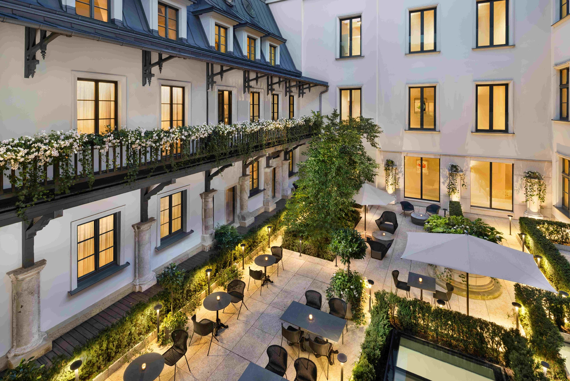 Illuminated courtyard with dining tables, black chairs, umbrellas, lush greenery, and white flowered balcony railings at dusk.