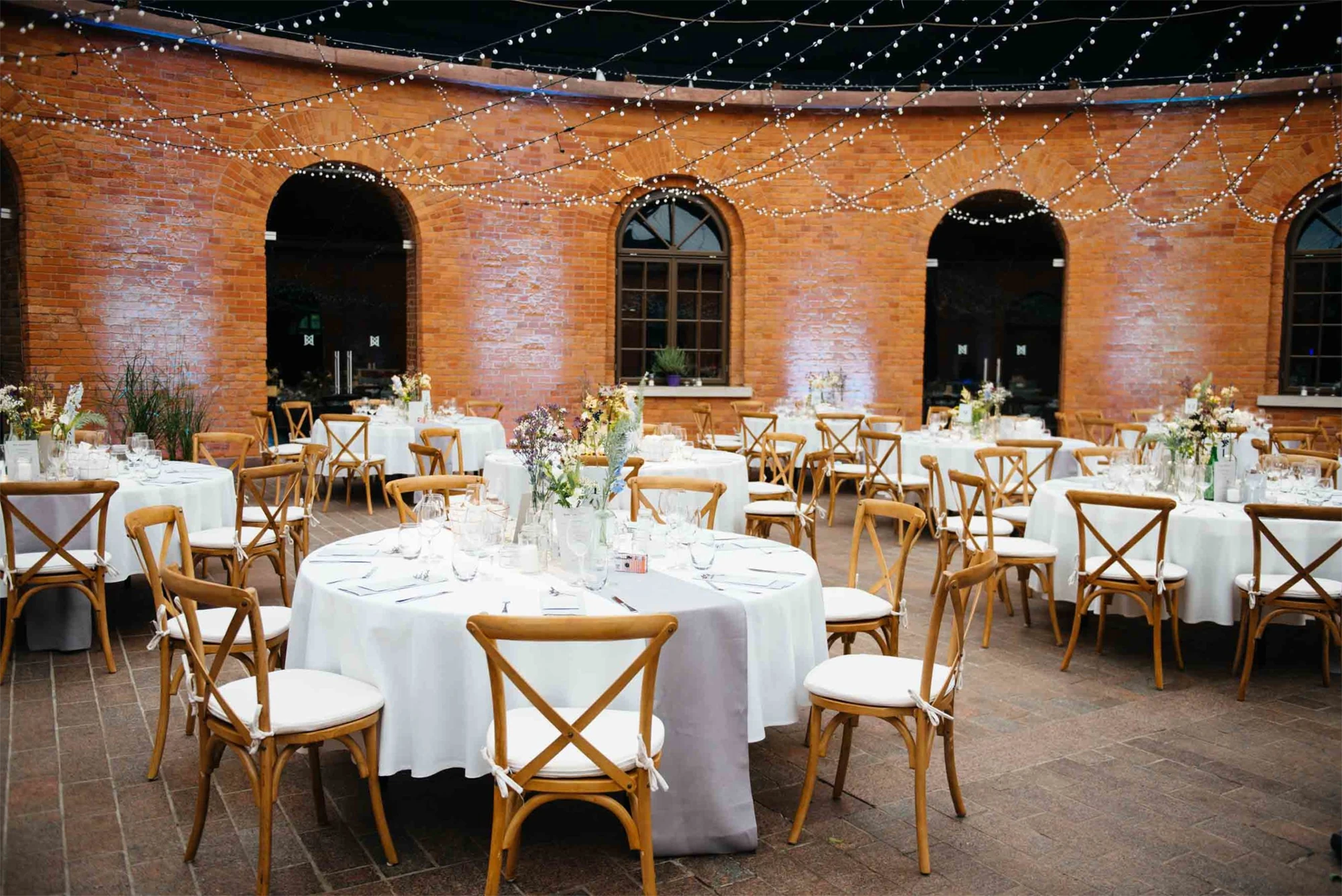 Elegant outdoor dining area with round tables set with white tablecloths, wooden chairs, and string lights overhead against a brick wall.