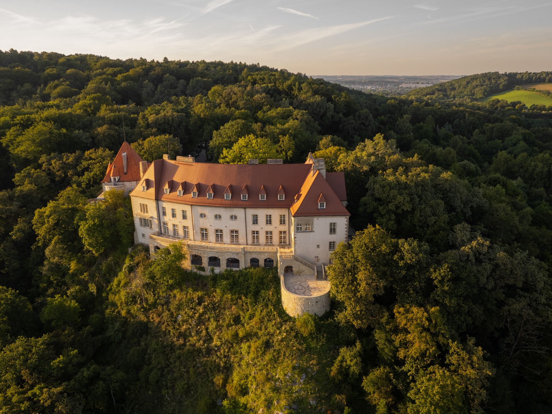 A large castle with a red roof perched on a forested hill under a soft evening sky.