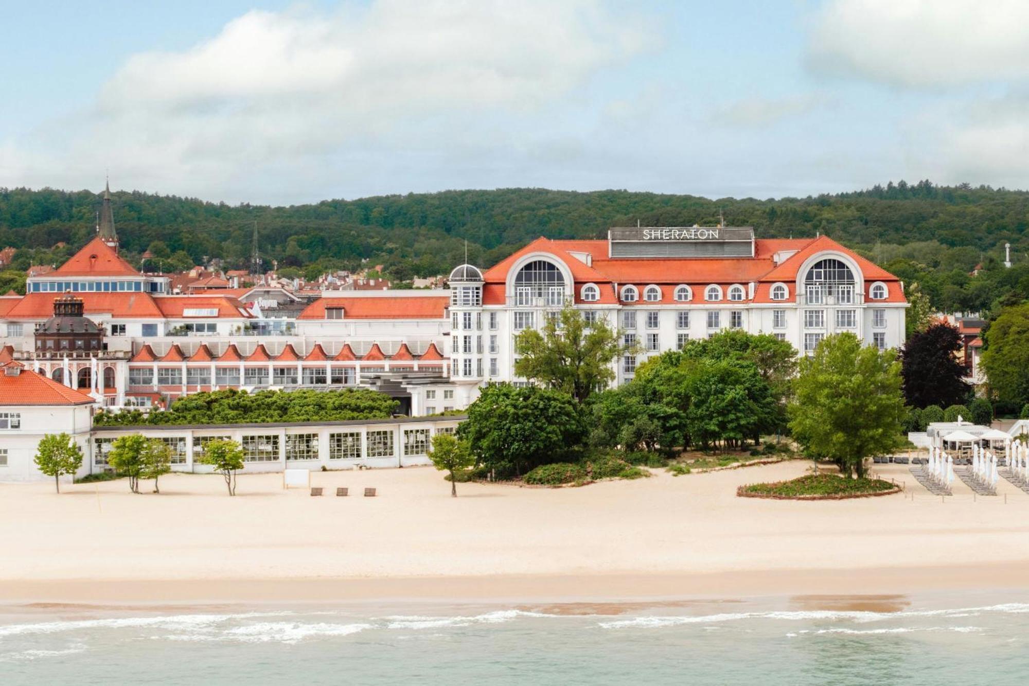 Sheraton hotel with red roofs facing a sandy beach and green trees under a partly cloudy sky.