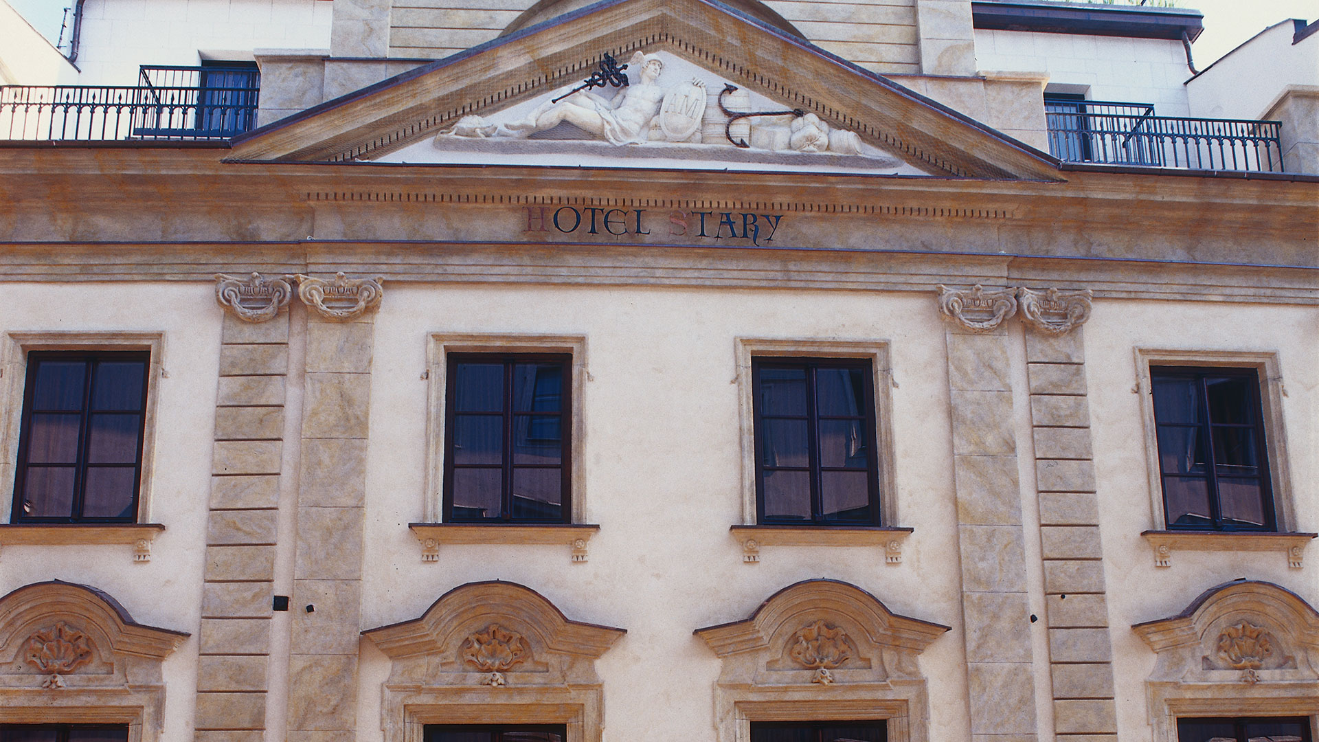 Facade of a classical-style building with four windows and a triangular pediment featuring a reclining figure sculpture and the text 'HOTEL STARY'.