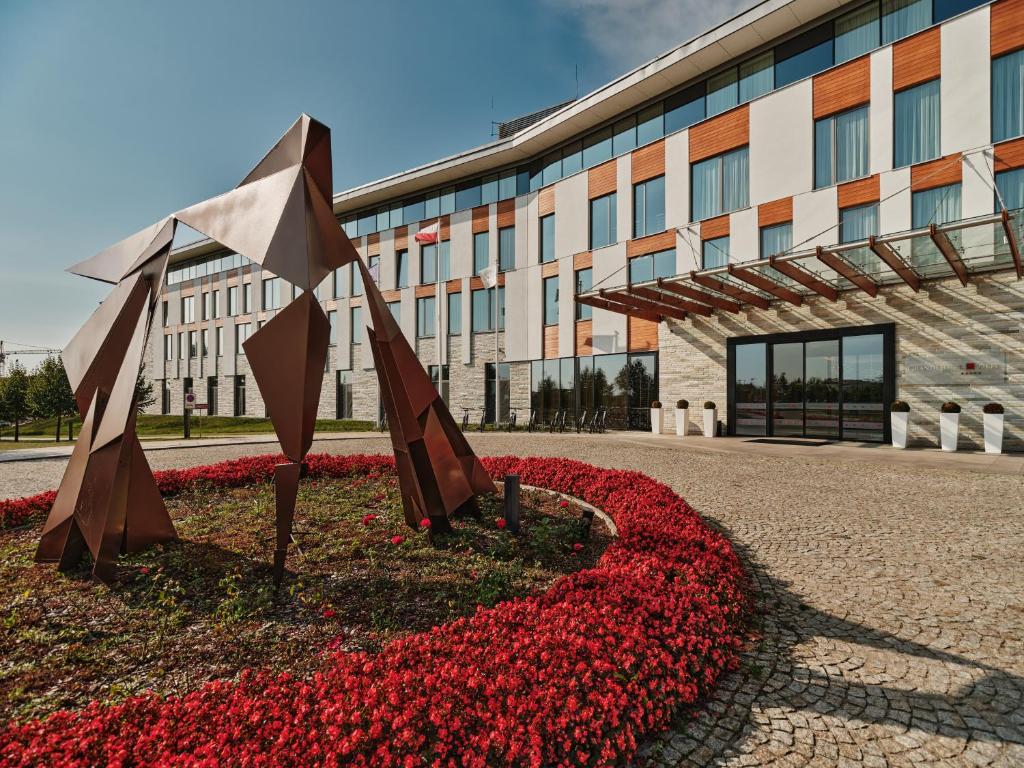 Luxurious bedroom with dark tufted headboard, wooden panel walls, large patterned rug, two grey armchairs by a bright window, and a beige bench at the foot of the bed.