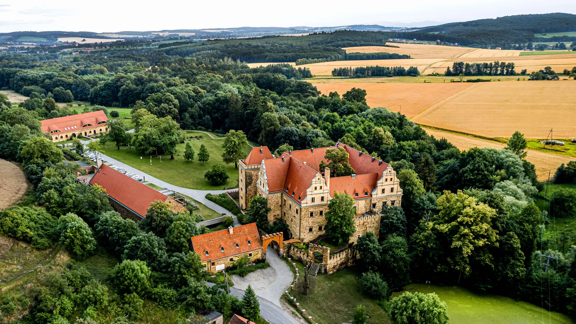Aerial view of a historic stone castle with red roofs surrounded by dense green forest and golden fields.