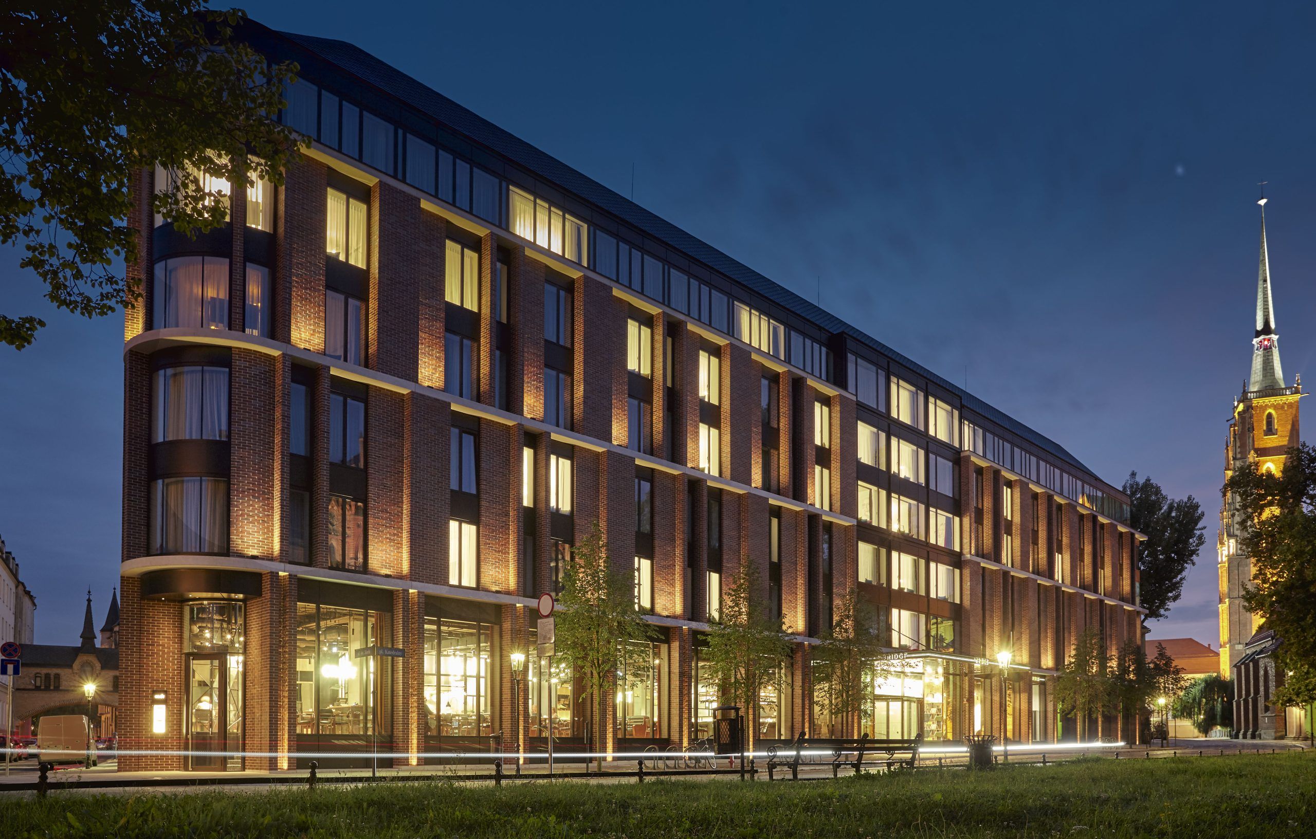 Modern brick building illuminated at dusk with a tall church spire in the background.