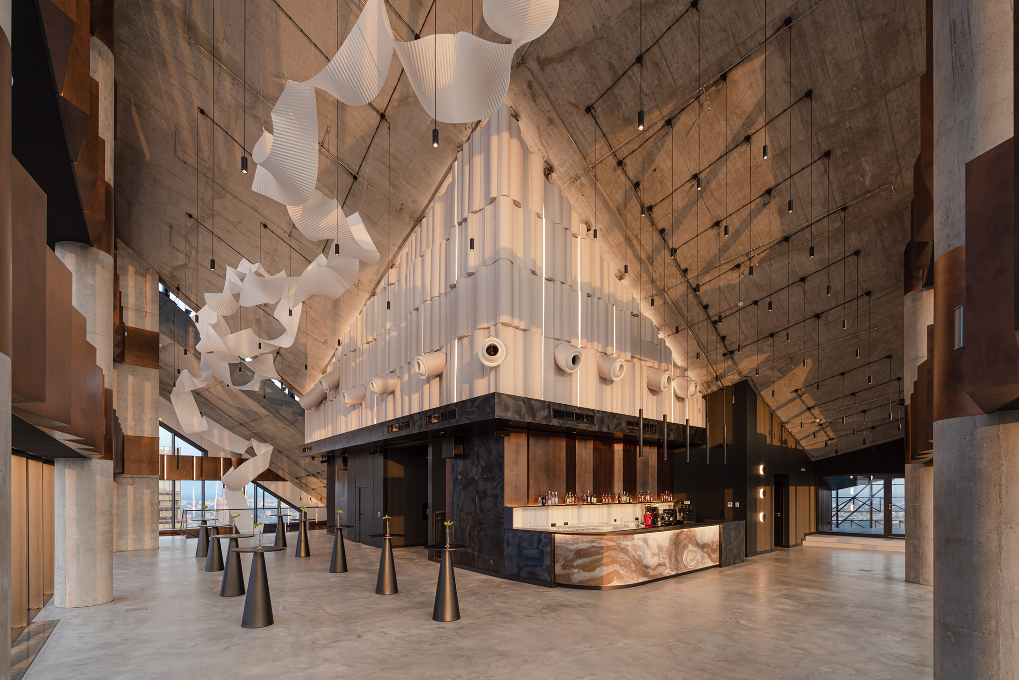 Modern bar area inside a large industrial-style building with high concrete ceilings, metal columns, sculptural white hanging decorations, and tall black cone-shaped tables with single flowers.