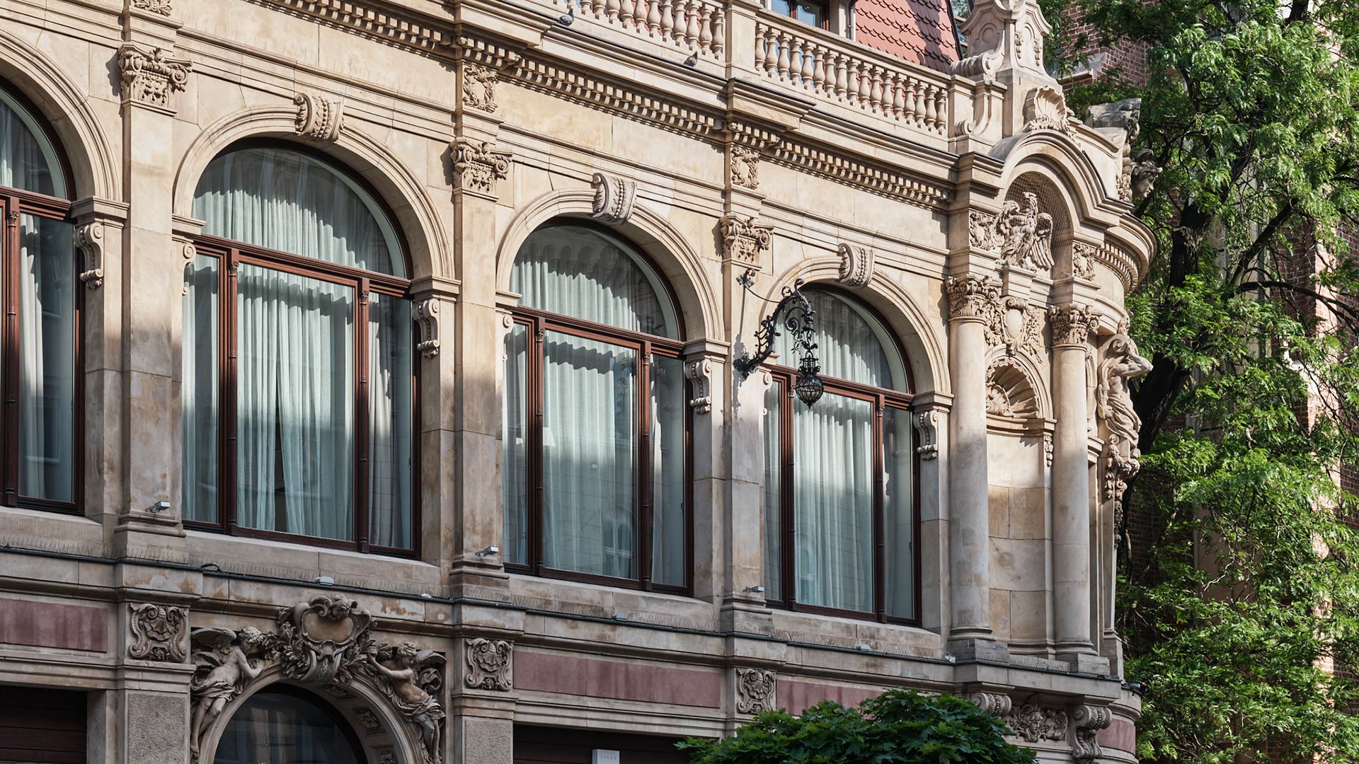 Historic building facade with large arched windows, ornate stone carvings, and green trees on the side.