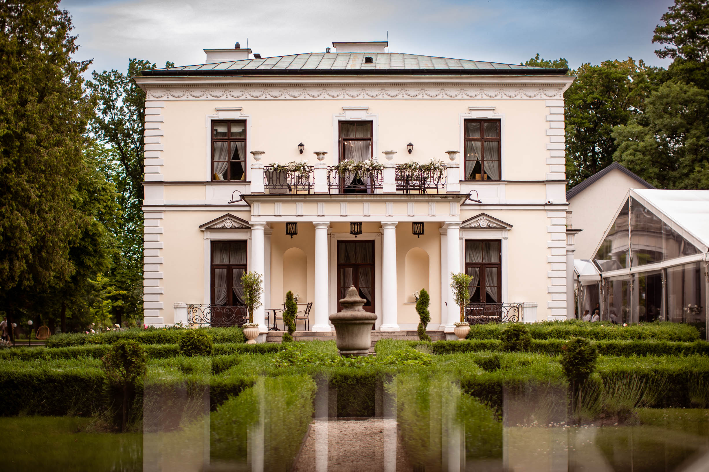 Elegant two-story cream-coloured mansion with white pillars, balcony with flowers, and manicured garden in front.