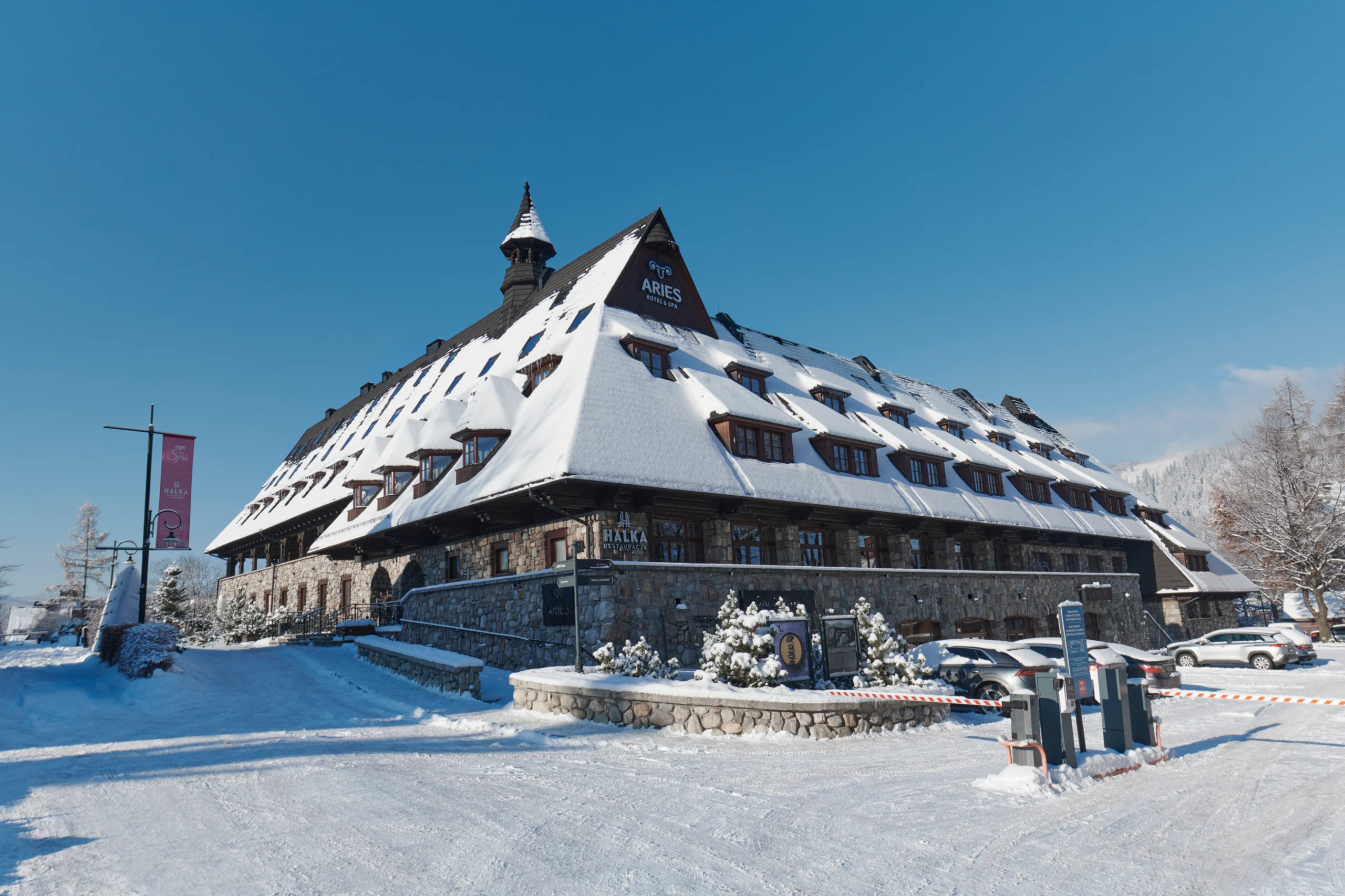 Snow-covered Aries Hotel & Spa with stone walls and steep roof under clear blue sky in a winter setting.