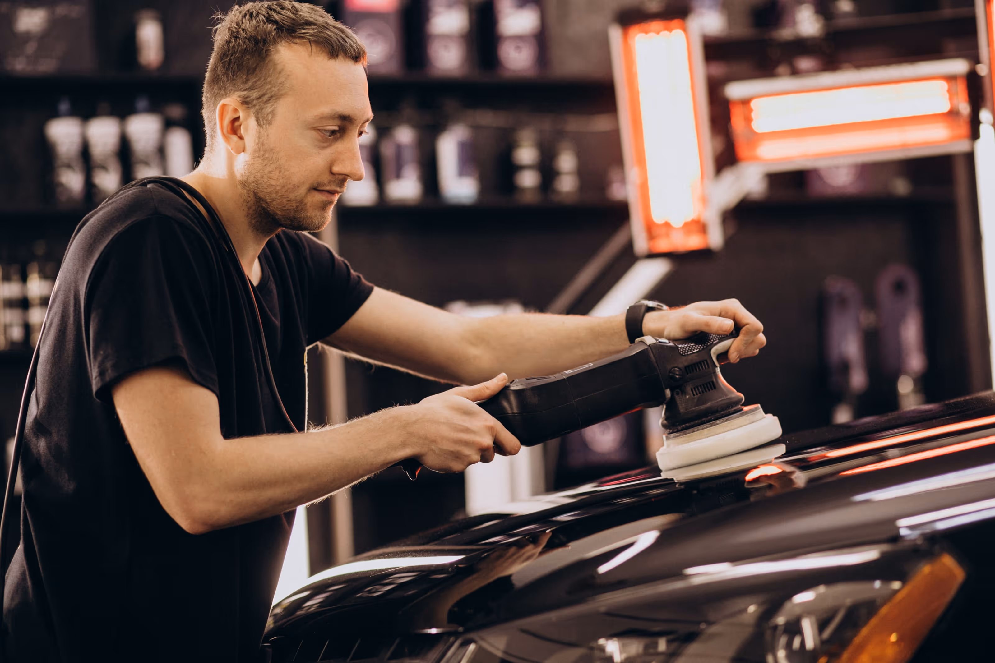 Homem polindo o capô de um carro preto com uma máquina de polimento em uma oficina de detalhamento automotivo.