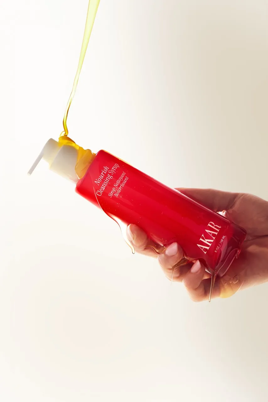 A hand holding Akar Nourish Cleansing Syrup bottle, dripping with golden liquid.
