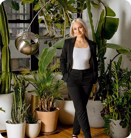 Blonde woman in black suit and white top standing among large green houseplants near a window.