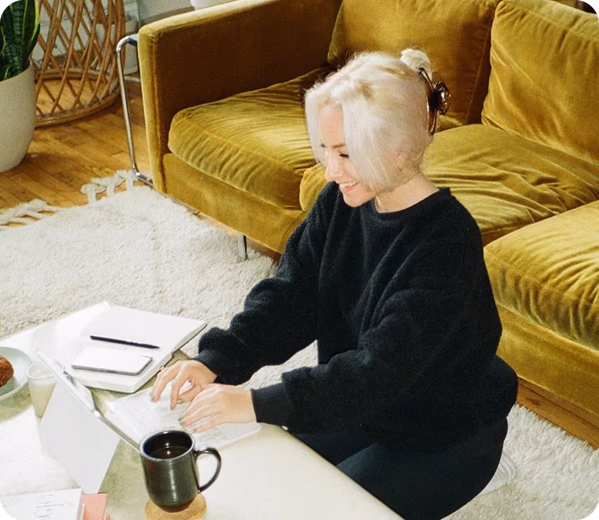 Smiling woman with tied up blonde hair, typing on a laptop while sitting on the floor in front of a mustard yellow couch.