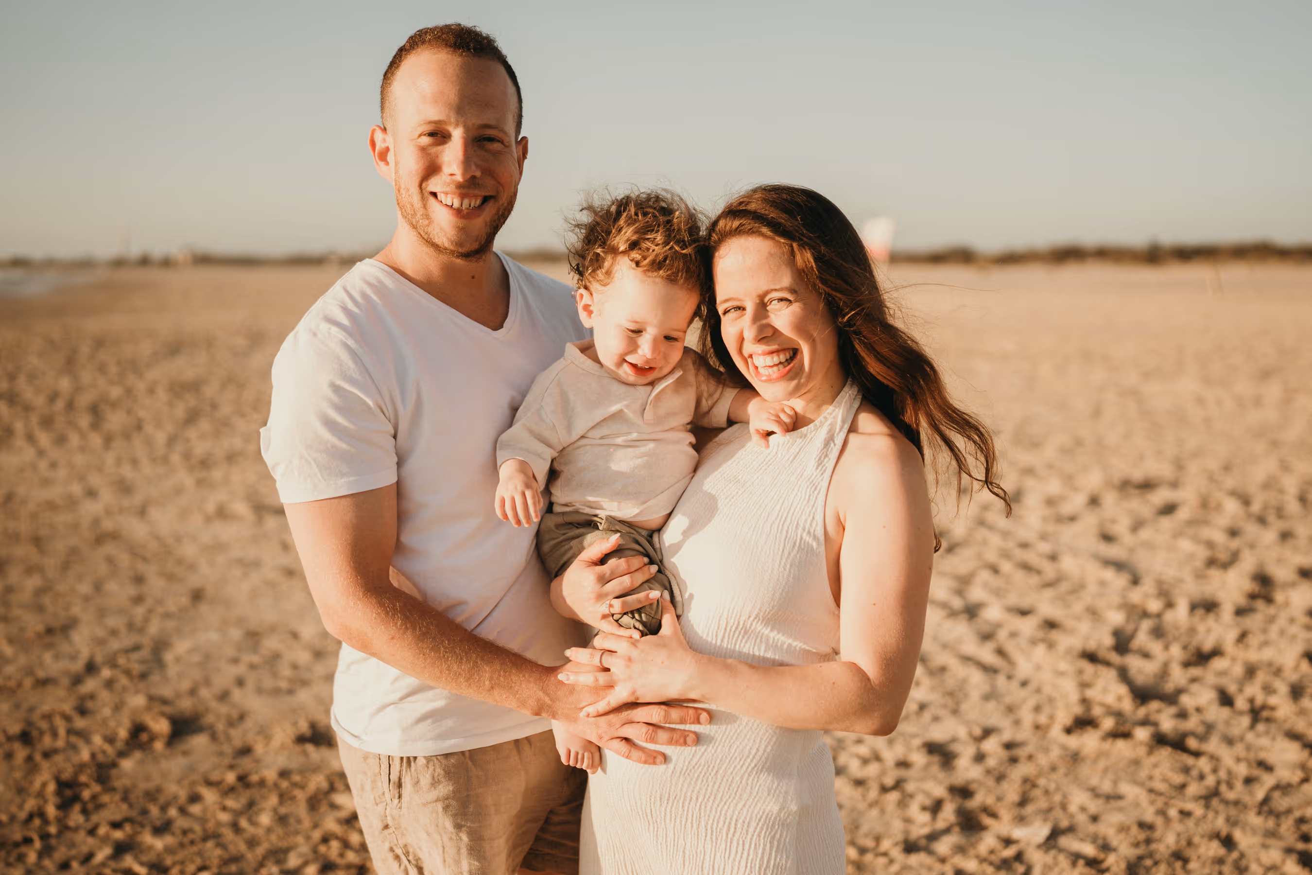 משפחה מחויכת עם אבא, אמא ותינוק בחוף ים בשקיעה.