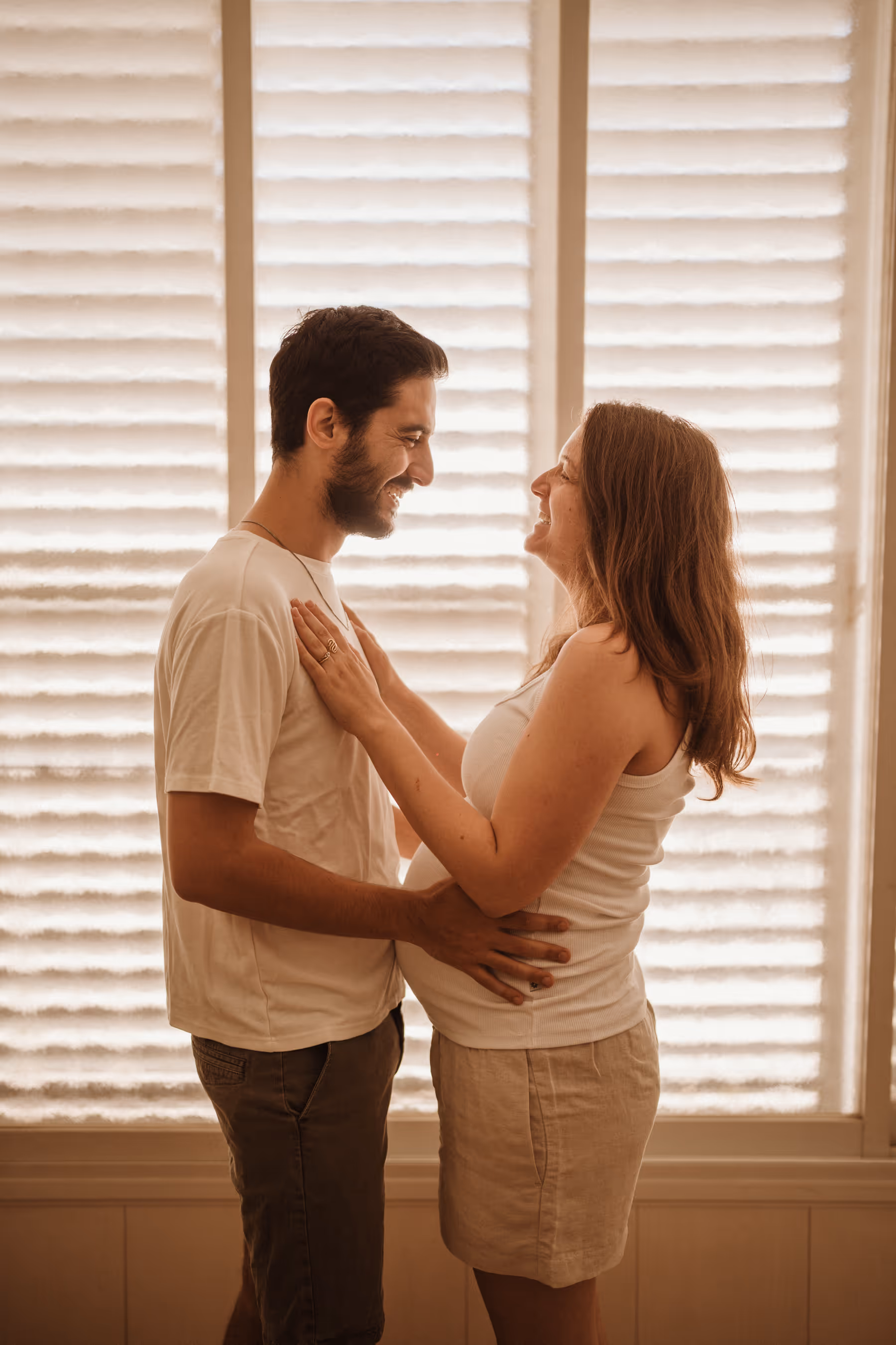 זוג עומד פנים מול פנים, האיש מחבק את האישה ההרה ומחייכים זה לזה.