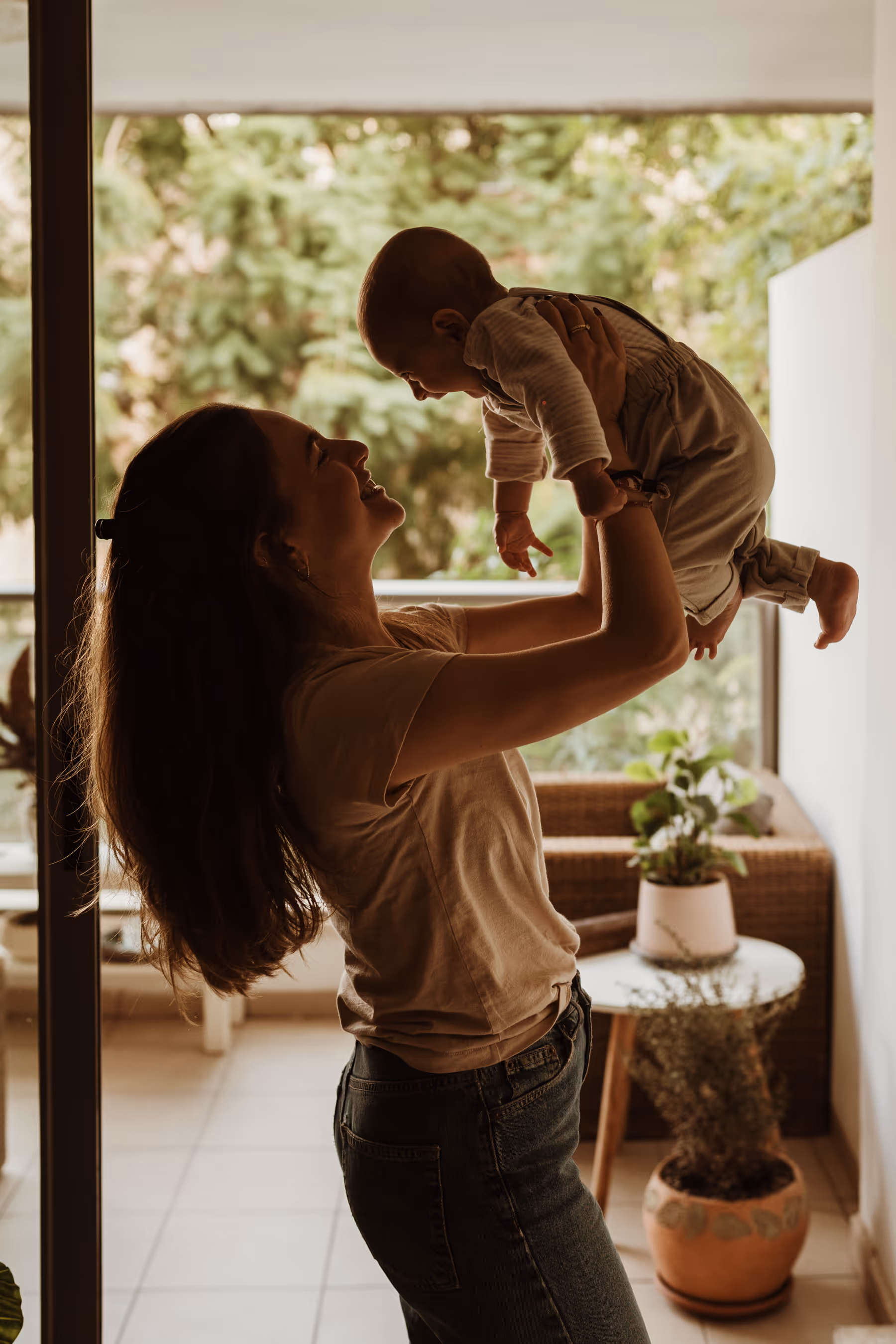 אישה מחזיקה תינוק באוויר בחיוך, ברקע מרפסת עם צמחים.