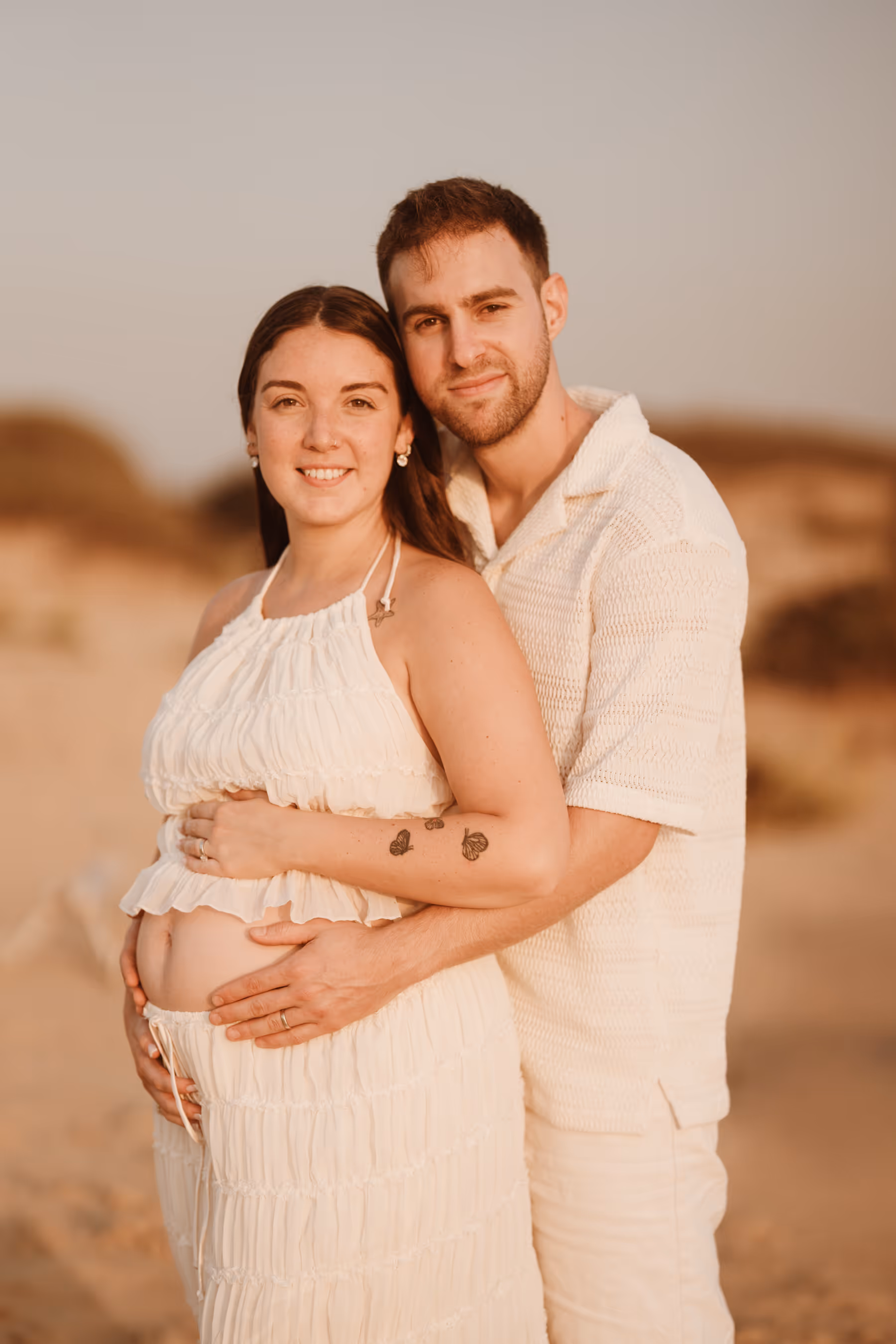 זוג מחובק בחוף ים, האישה בהריון ולובשת שמלה לבנה, השניים מחייכים למצלמה בשעת שקיעה.