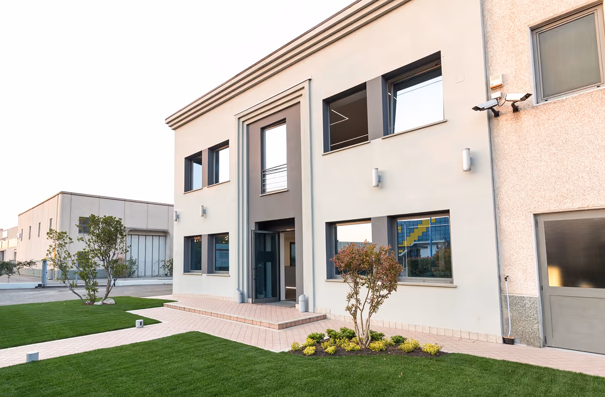 Edificio commerciale moderno con ingresso centrale, finestre ampie e giardino ben curato con prato verde e piante.