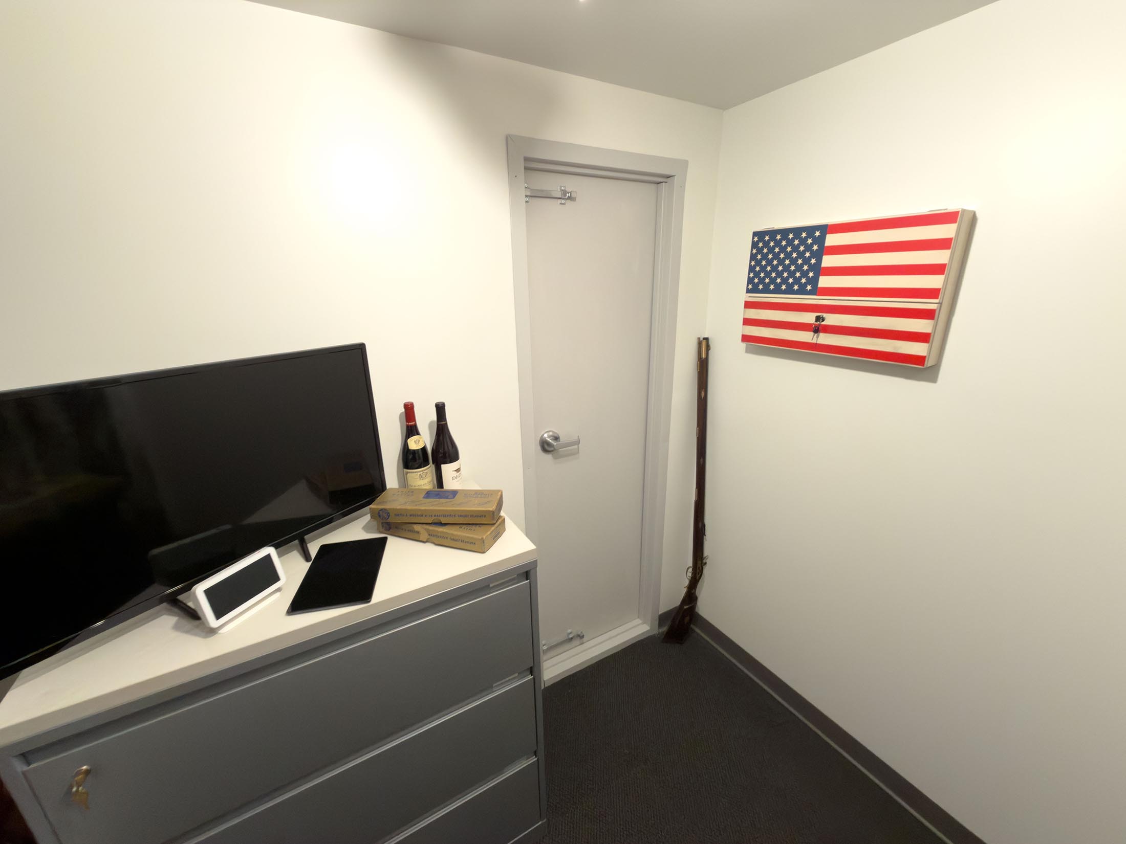 Corner of an office with a gray filing cabinet holding a TV, digital clock, tablet, two wine bottles, and two pizza boxes; a closed gray door; a rifle leaning against the wall; an American flag box with keys hanging on the wall.