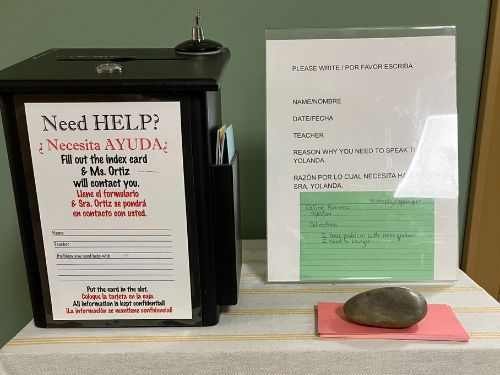 Black help box with instructions in English and Spanish to fill out an index card for contact, next to a clear stand holding bilingual form sheets on a table with a rock paperweight.