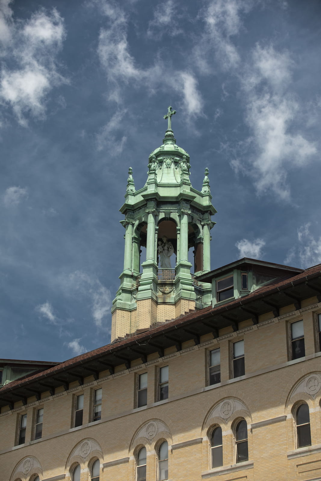 Green copper church steeple with a cross atop and a statue inside, set against a blue sky with clouds.