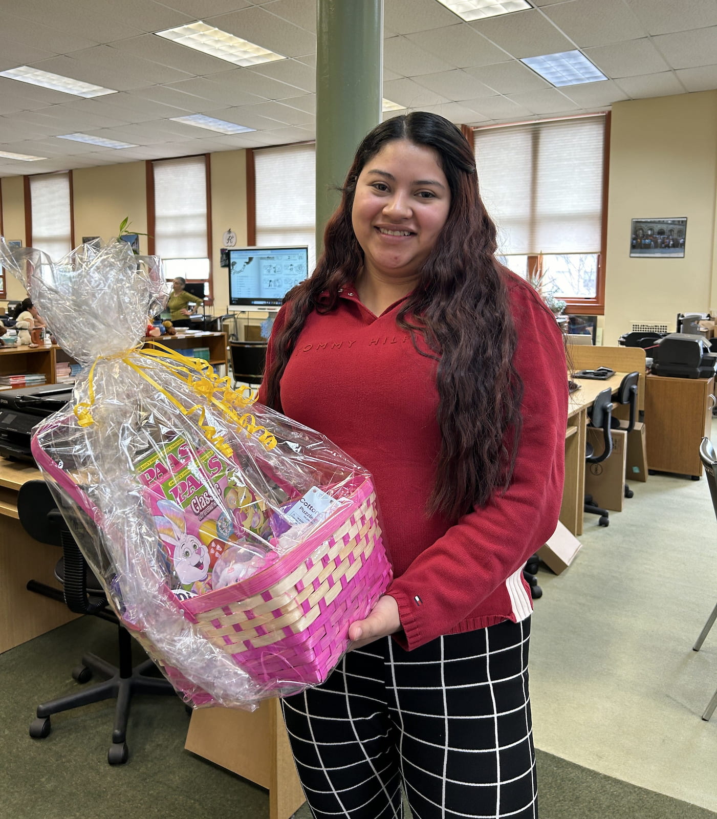 Smiling woman in a red sweater holding a pink woven gift basket wrapped with clear plastic and yellow ribbon in an office setting.