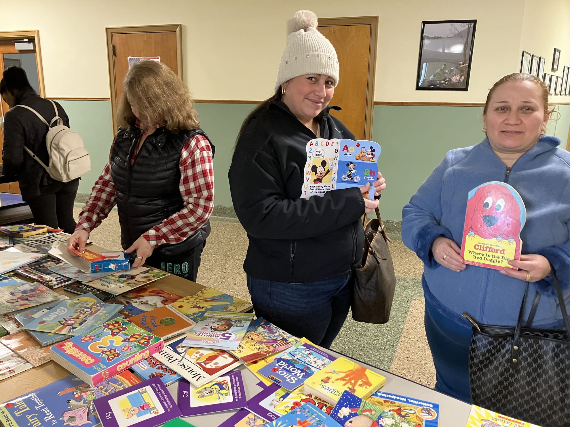 Three women at a book donation table holding children’s books, with many colorful books spread out on the table in front of them.