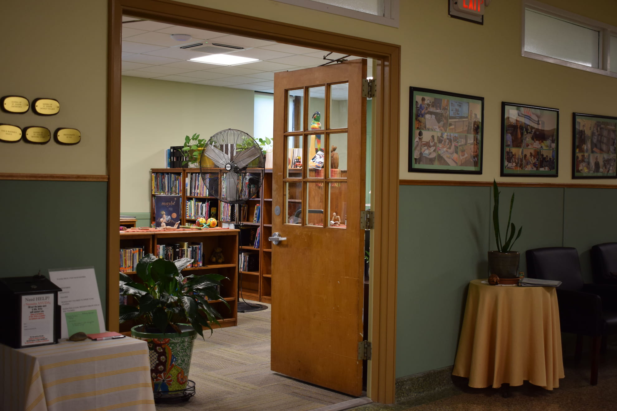 Open wooden door leading to a library room with bookshelves, a large fan, and a colorful parrot decoration; hallway with framed photos and plants on tables.