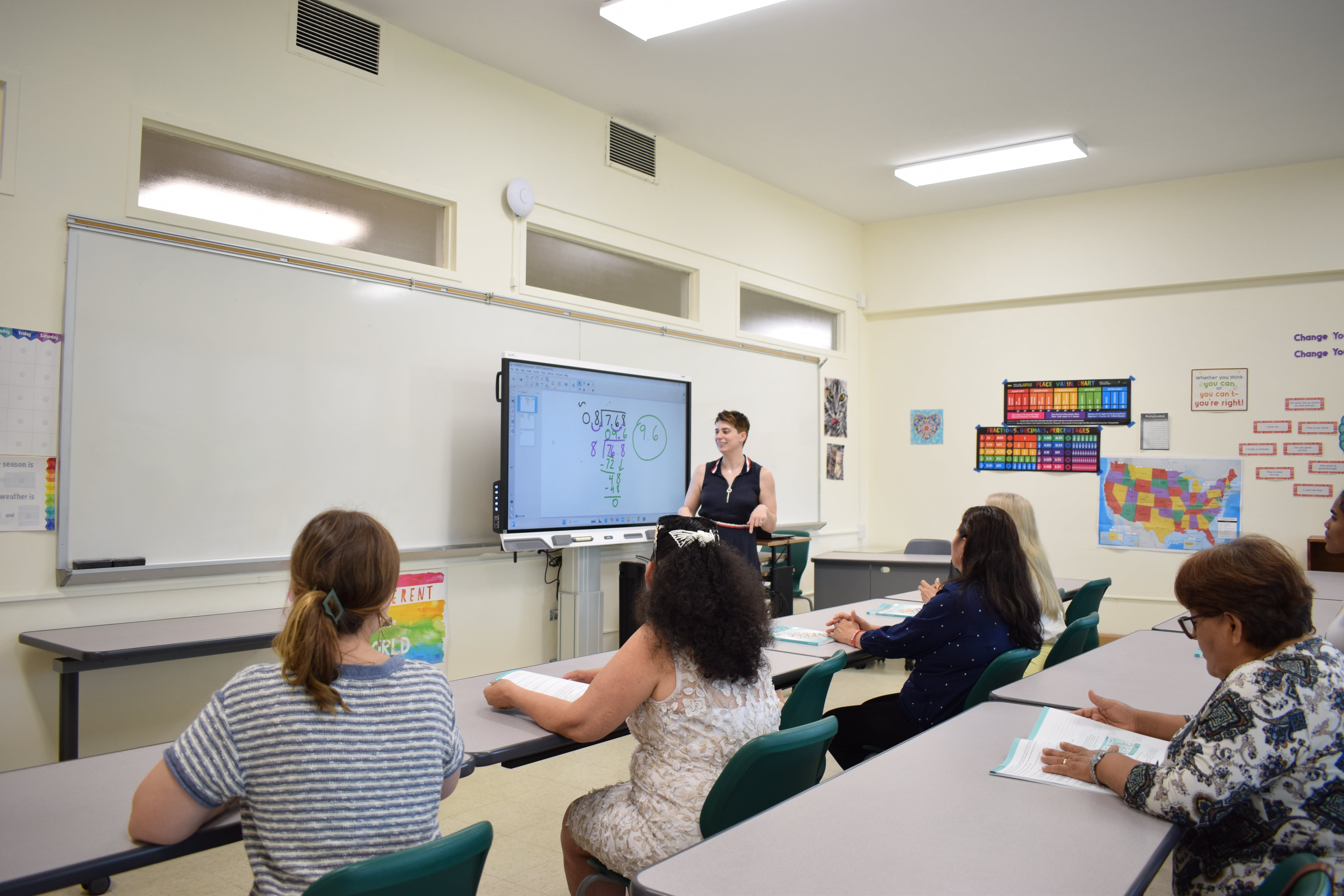 Students seen from behind, looking at the classroom board while the teacher explains a lesson.