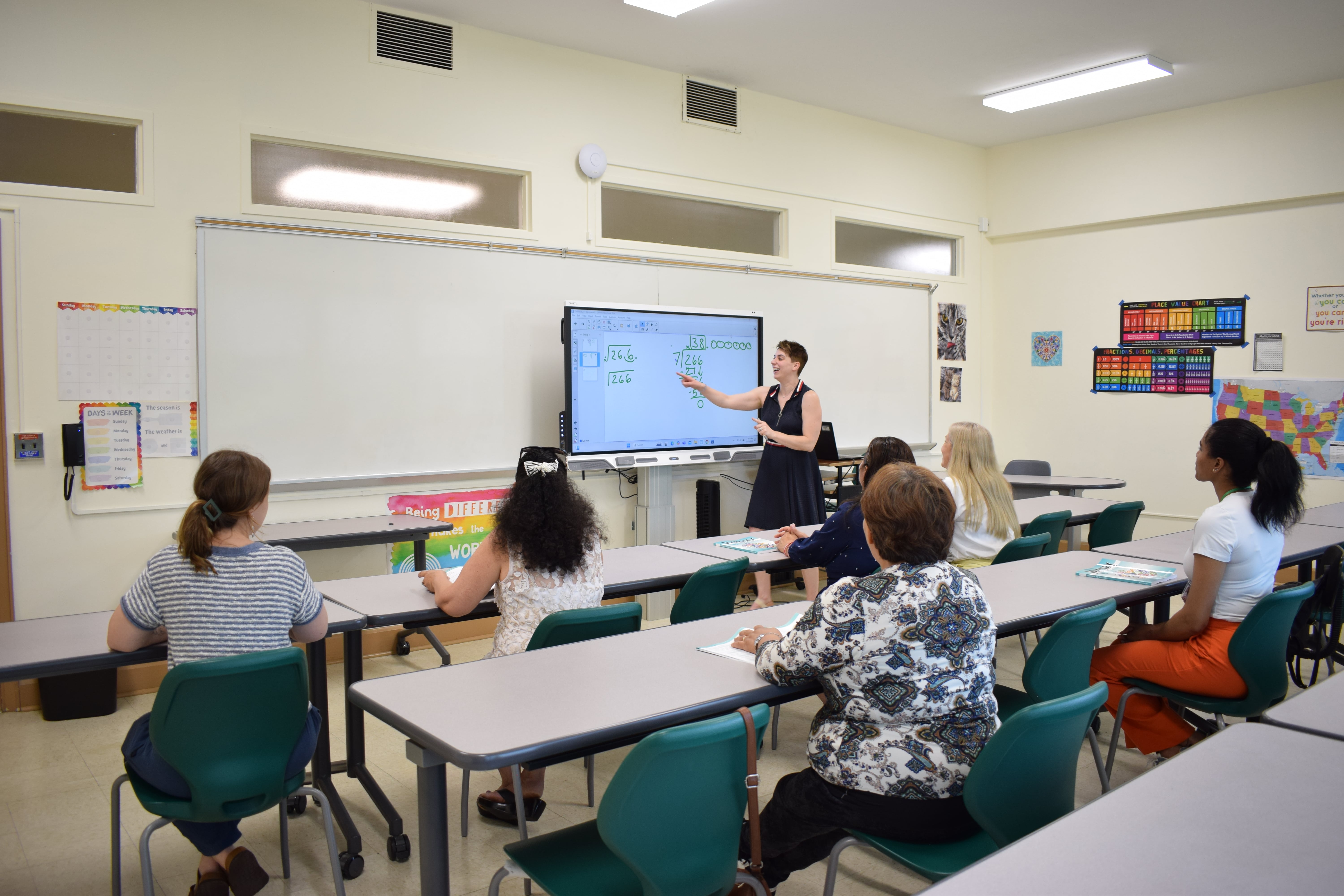 Students seen from behind, looking at the classroom board while the teacher explains a lesson.