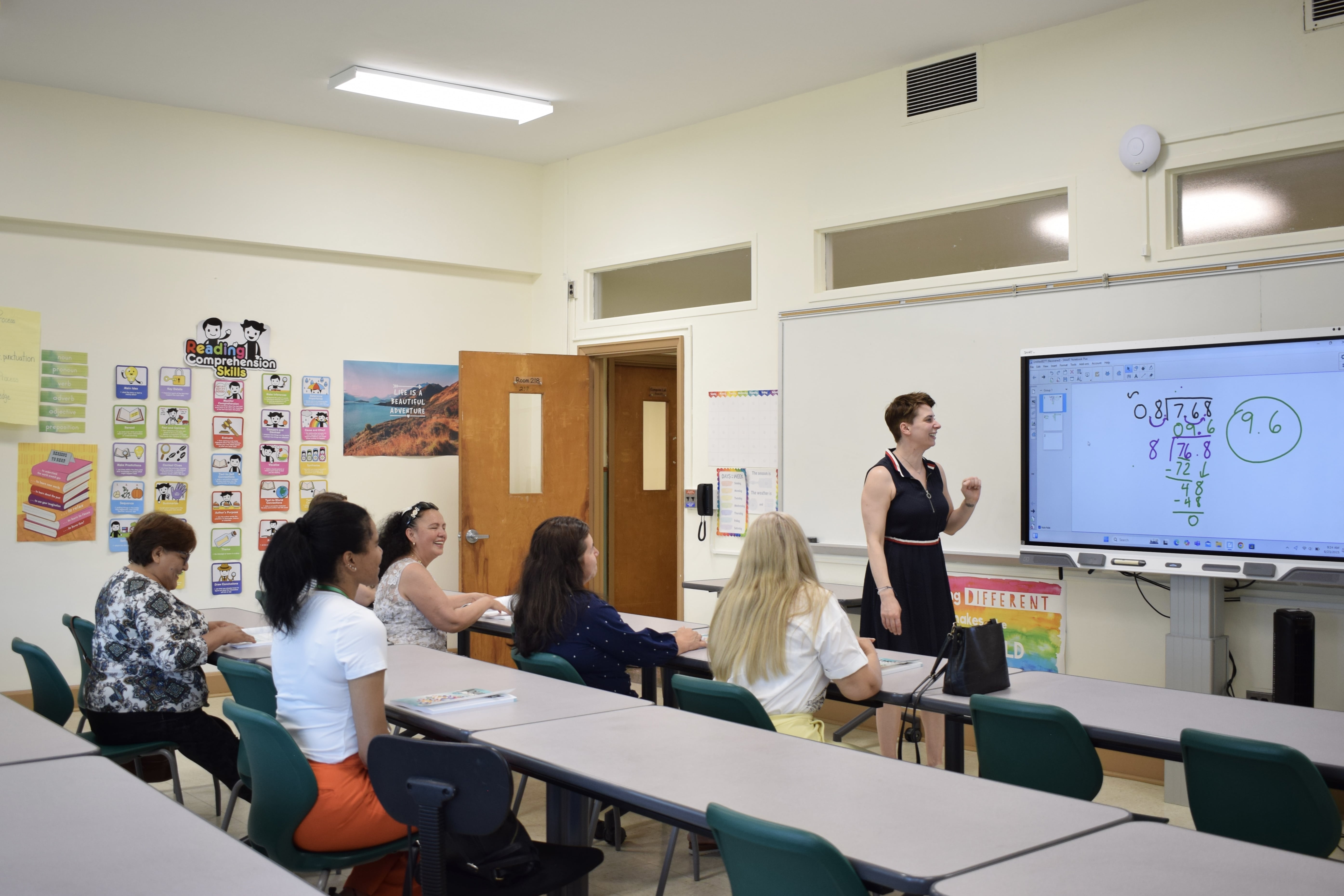 Students seen from behind, looking at the classroom board while the teacher explains a lesson.