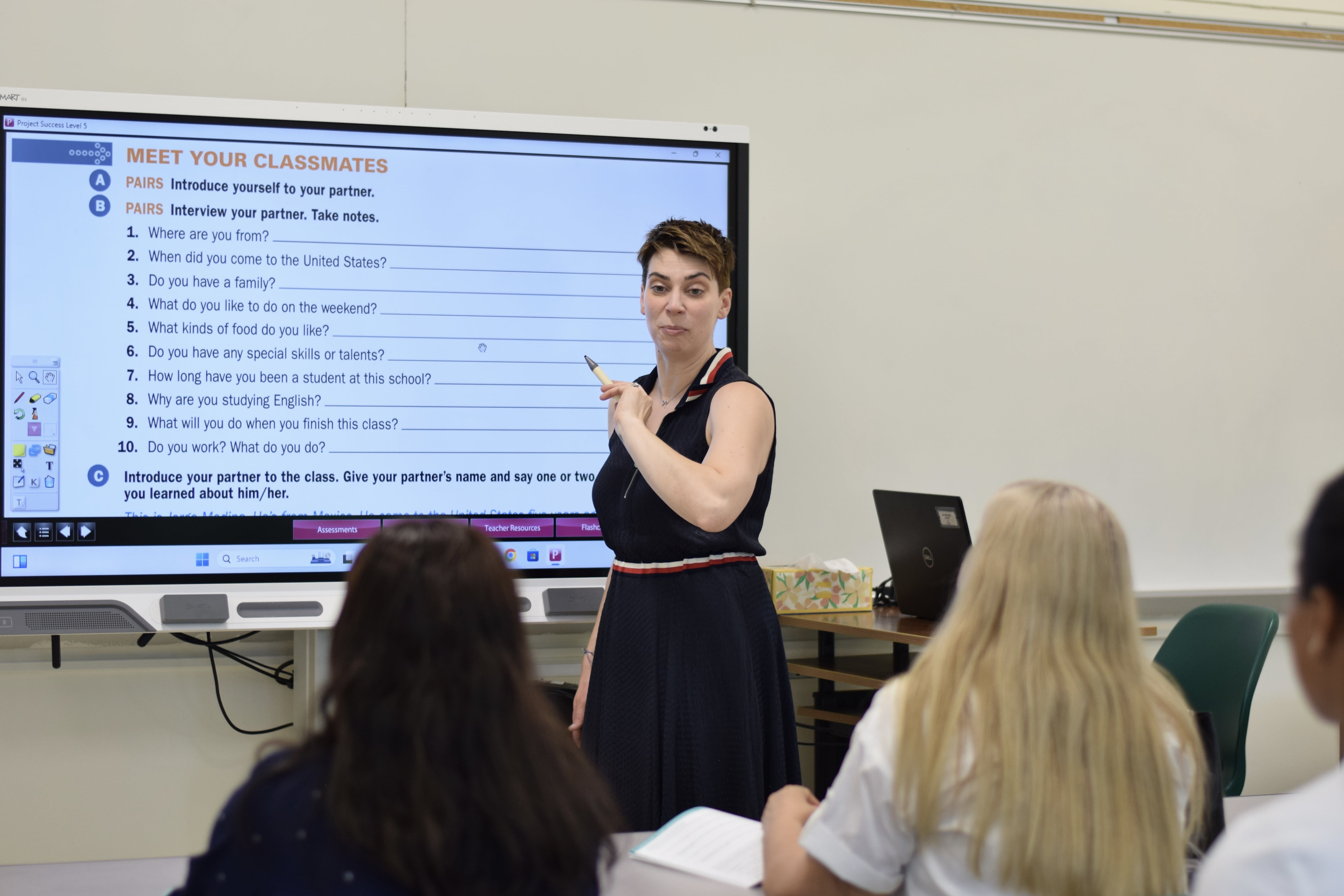 Students seen from behind, looking at the classroom board while the teacher explains a lesson.