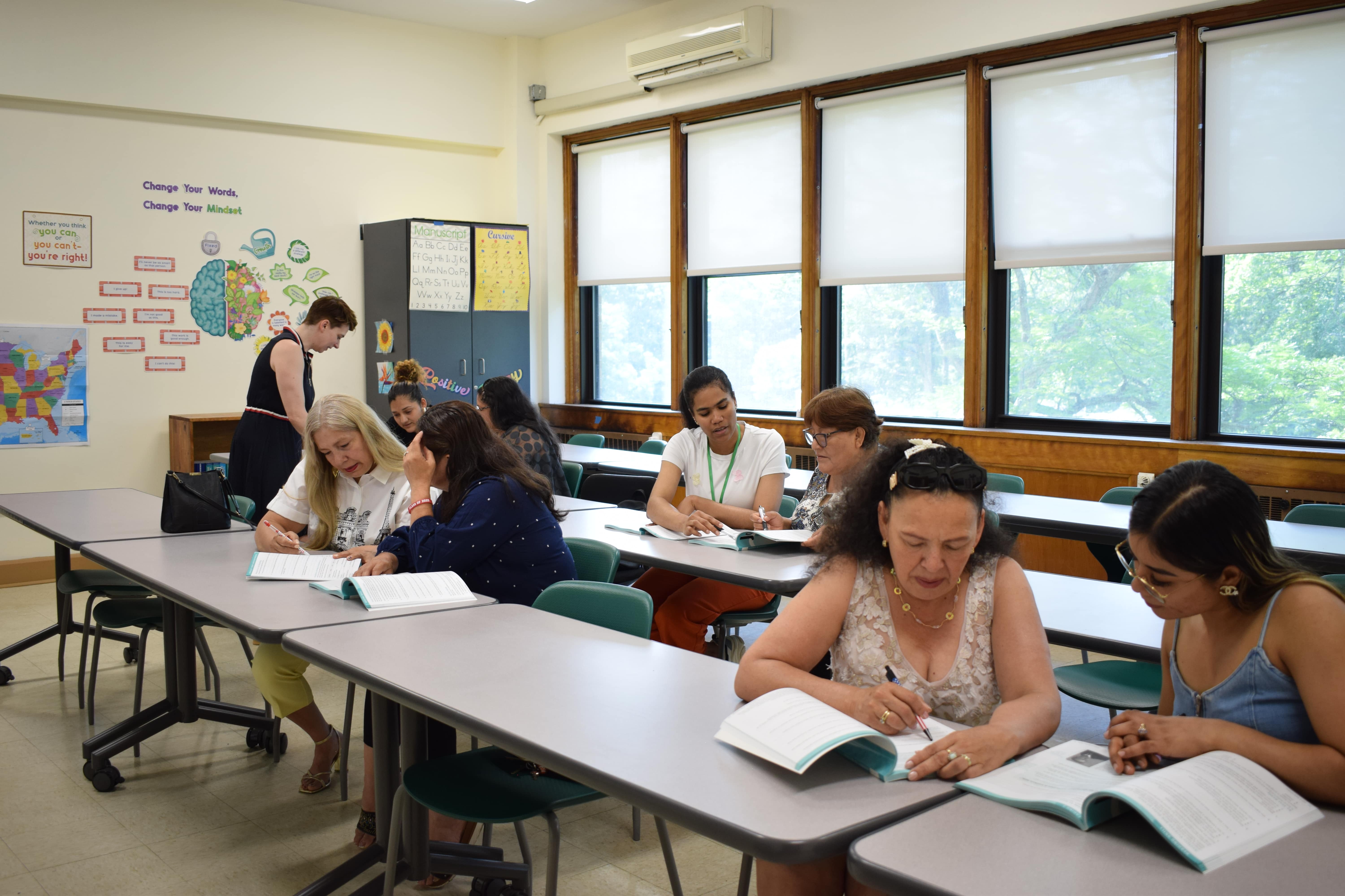 Students sit in a classroom reading from books while the teacher walks around and assists them.